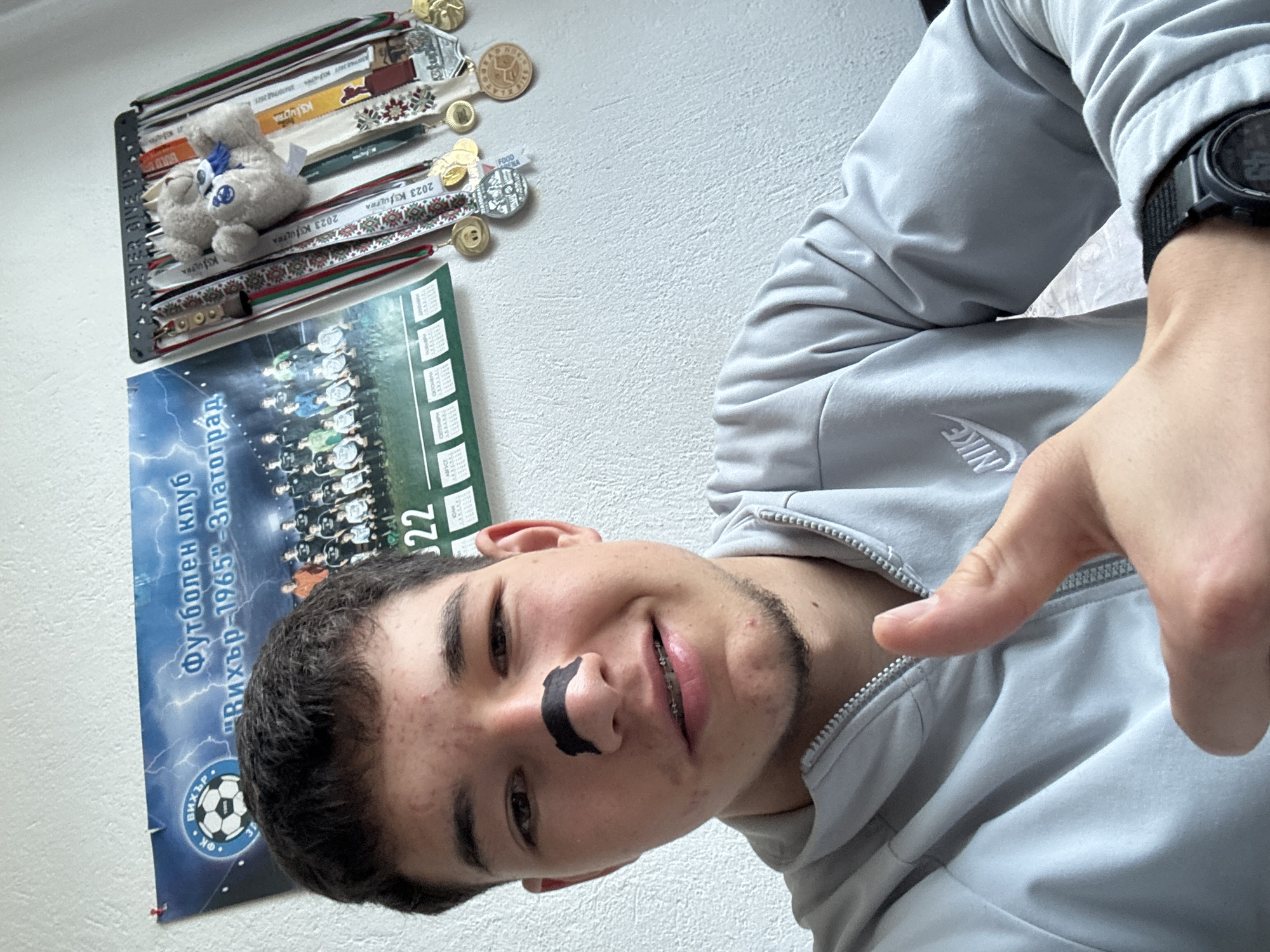 A selfie of a young man with a black adhesive strip across his nose. He has braces and is wearing a gray Nike hoodie, pointing towards his chest with his right hand, on which he is wearing a digital watch. Behind him is a white wall featuring a shelf holding medals and a stuffed animal, along with a poster for the "Вихър-1965" football club from Zlatograd, Bulgaria.