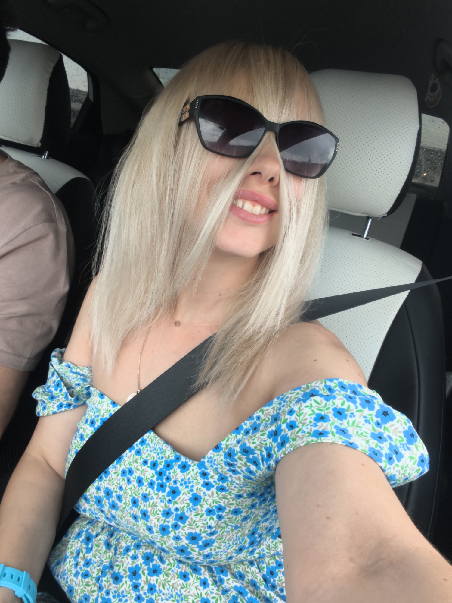 A young woman with blonde hair and sunglasses is seated in the passenger seat of a car. She is wearing a blue floral dress and a seatbelt. Her hair partially obscures her face, but she is smiling. The interior of the car is visible, including the headrest of the driver's seat. The background appears to be a rainy day. The image is a selfie taken from the perspective of the woman.