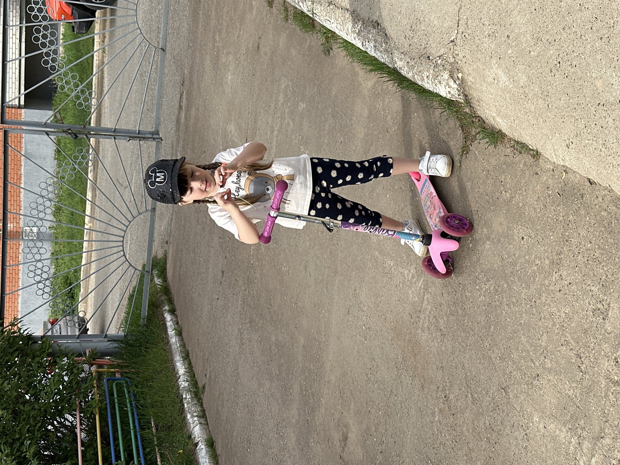 A young girl with braided hair stands on a pink scooter on a concrete surface, holding the scooter's handlebars. She wears a black baseball cap with a white "M" logo, a white t-shirt with a cartoon bear print, and black leggings with a white floral pattern. She is smiling and looking at the camera.
In the background, there is a metal fence and a brick building. There is also grass and some bushes visible. The location is the city of Chaikovsky, Russia.