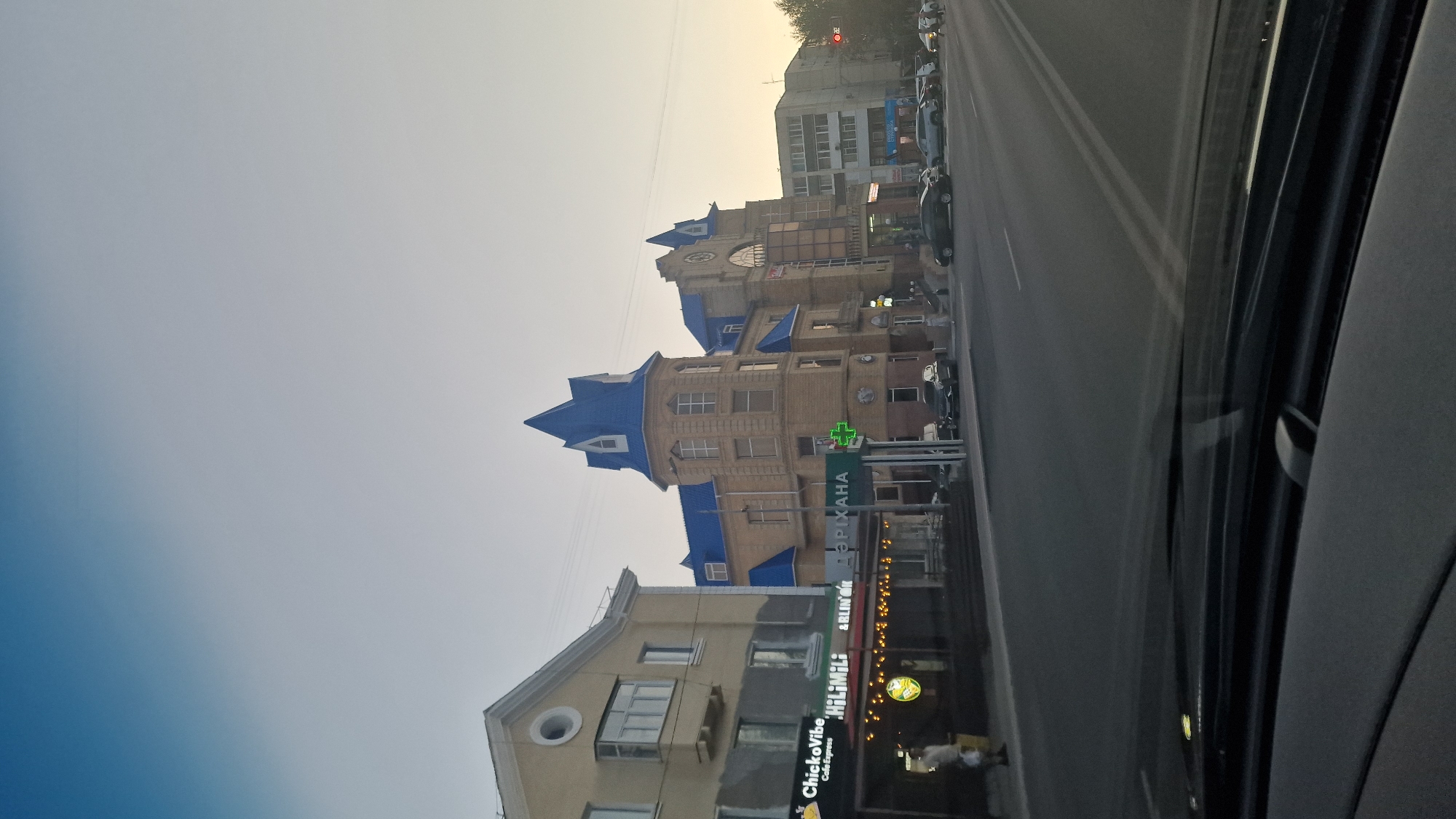 The image depicts a street scene in Kostanay, Kazakhstan, at what appears to be dusk or early morning. The focal point is a distinctive building with a blue, conical roof and elaborate architectural details, likely a historical or public building. To its left is another building with a lighter facade and a bay window. Below these buildings, a row of businesses line the street, including a cafe named "ChickeVibe" and another with signage that reads "HiHiMili". A green cross symbol, indicating a pharmacy, is visible on a pole in front of the building with the blue roof. The street in the foreground is paved and appears to be a multi-lane road with vehicles, including cars and SUVs, parked or driving. The sky above is a gradient of light blue and grey, transitioning towards the horizon. The overall impression is a calm urban environment on a street with commercial establishments and residential buildings.