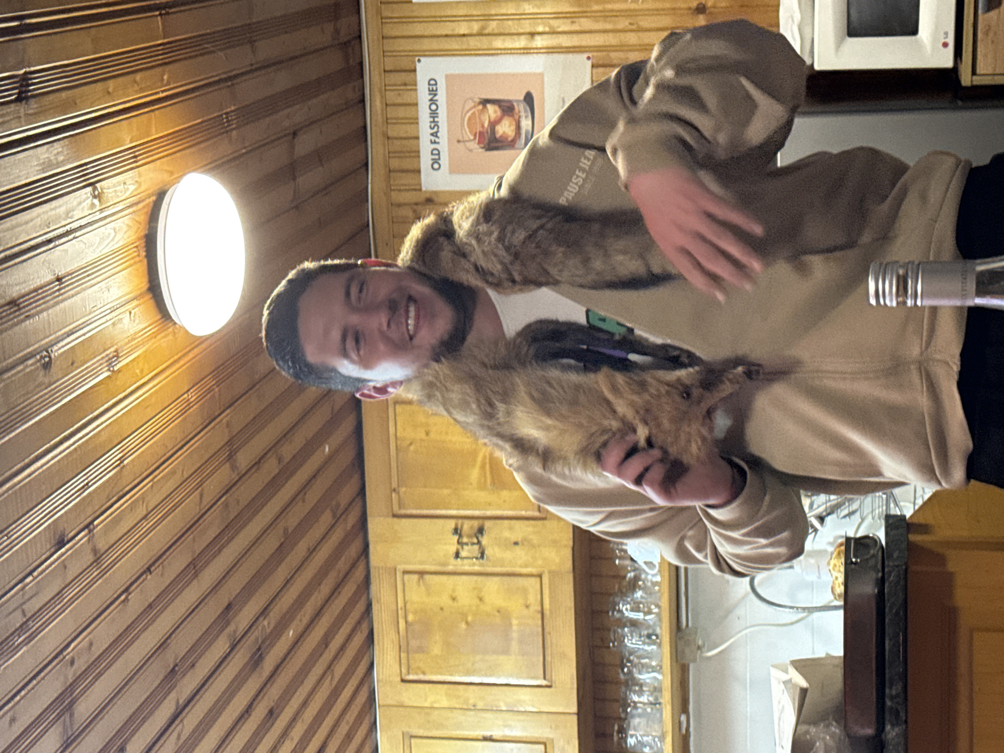 A man smiles while holding a fur stole in a room with wood paneling. A round light fixture is attached to the wall. A poster reading "Old Fashioned" is also on the wall, above wooden cabinets. A microwave is visible above a counter. There is no information in the image that places this scene in Lilkovo, Bulgaria.