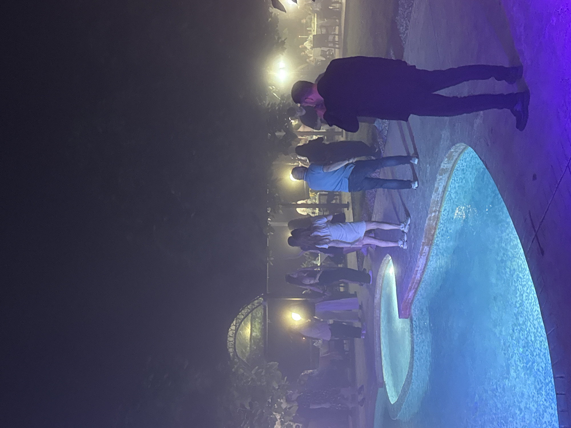 The image depicts an outdoor nighttime social event in Sredets, Bulgaria. A kidney-shaped swimming pool, illuminated with blue light, occupies the lower left and central foreground. The pool's edge is bordered by a paved surface, which transitions to a concrete or stone patio. In the immediate foreground to the right, a male subject is viewed from behind, wearing dark attire (jacket, pants, shoes), holding an object to his mouth, possibly a microphone or a drink.
Multiple individuals are distributed across the mid-ground and background. Near the pool's edge in the mid-ground, a male subject in a blue polo shirt and jeans stands, facing right. Several other people are standing and mingling in the mid-ground, including a woman in a light-colored dress facing away from the camera. The scene is characterized by a hazy or misty atmosphere, diffusing light sources and creating glowing halos around them. Bright lights are visible throughout the background, illuminating clusters of people. In the distant background, several groups are seated at tables with white tablecloths and chairs, suggesting a dining area. An arched, possibly decorative, structure is discernible in the left-center background. Overhanging foliage and trees are visible in the mid-ground and background. The ground in the foreground right exhibits a faint purple hue, likely from ambient lighting.