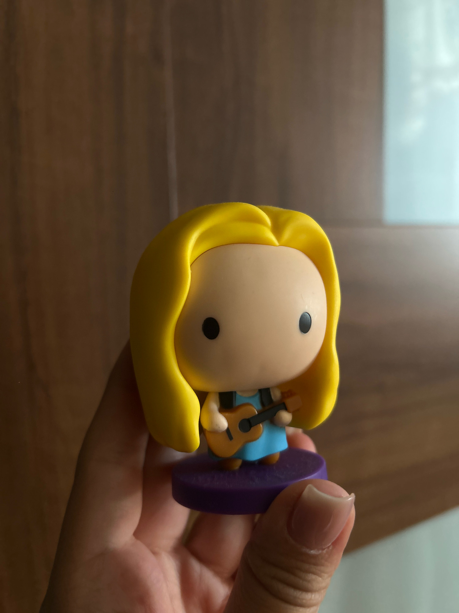 A person's hand holds a small, stylized figurine with blonde hair, a blue dress, and a guitar. The figurine is positioned on a purple base. The background consists of a wooden surface and a blurry, light-colored area. The image appears to be taken indoors, with natural lighting casting shadows. The location is not discernible from the image.