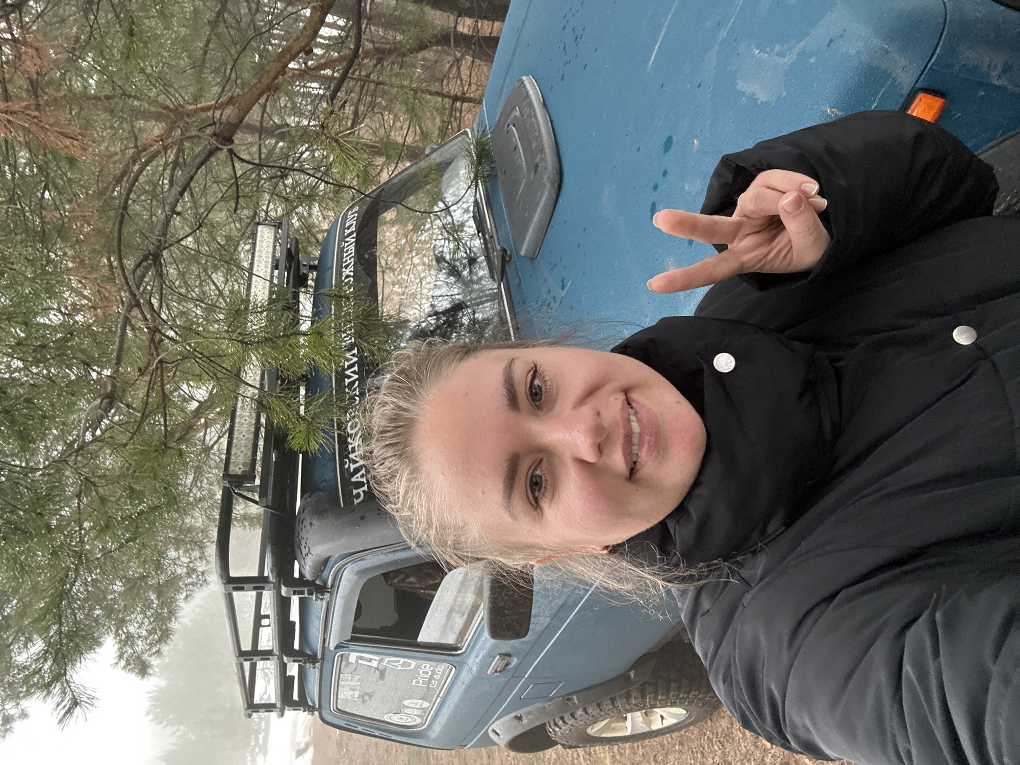 A young woman poses for a selfie outdoors in front of a blue off-road vehicle. She is wearing a black jacket and giving a peace sign with her right hand. The vehicle has a roof rack and additional lights. Text in Russian is visible on the windshield. The background includes a tree with thin branches, and a foggy or misty atmosphere suggesting a wooded or rural area. Based on the windshield text, the location is assumed to be in the city of Chaikovsky, Russia.