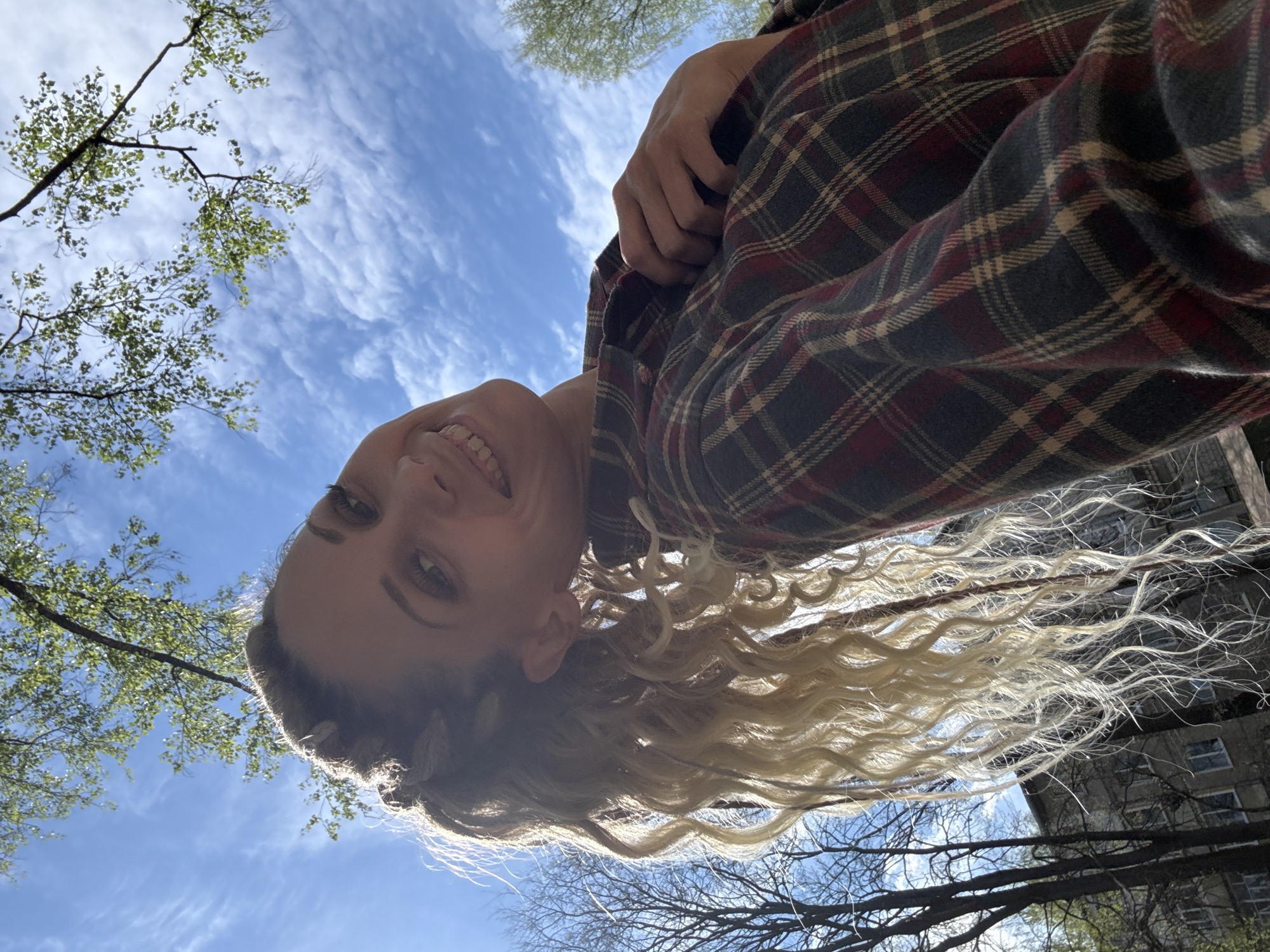 A low-angle shot looking up at a woman with blonde curly hair wearing a red and black plaid shirt. The background includes a bright blue sky with light white clouds and tree branches with some green leaves. A building is partially visible on the right edge of the frame. The woman is smiling and looking down at the camera. The location is likely Rusanovo, Russia, given the prompt, though no specific markers in the image confirm this.