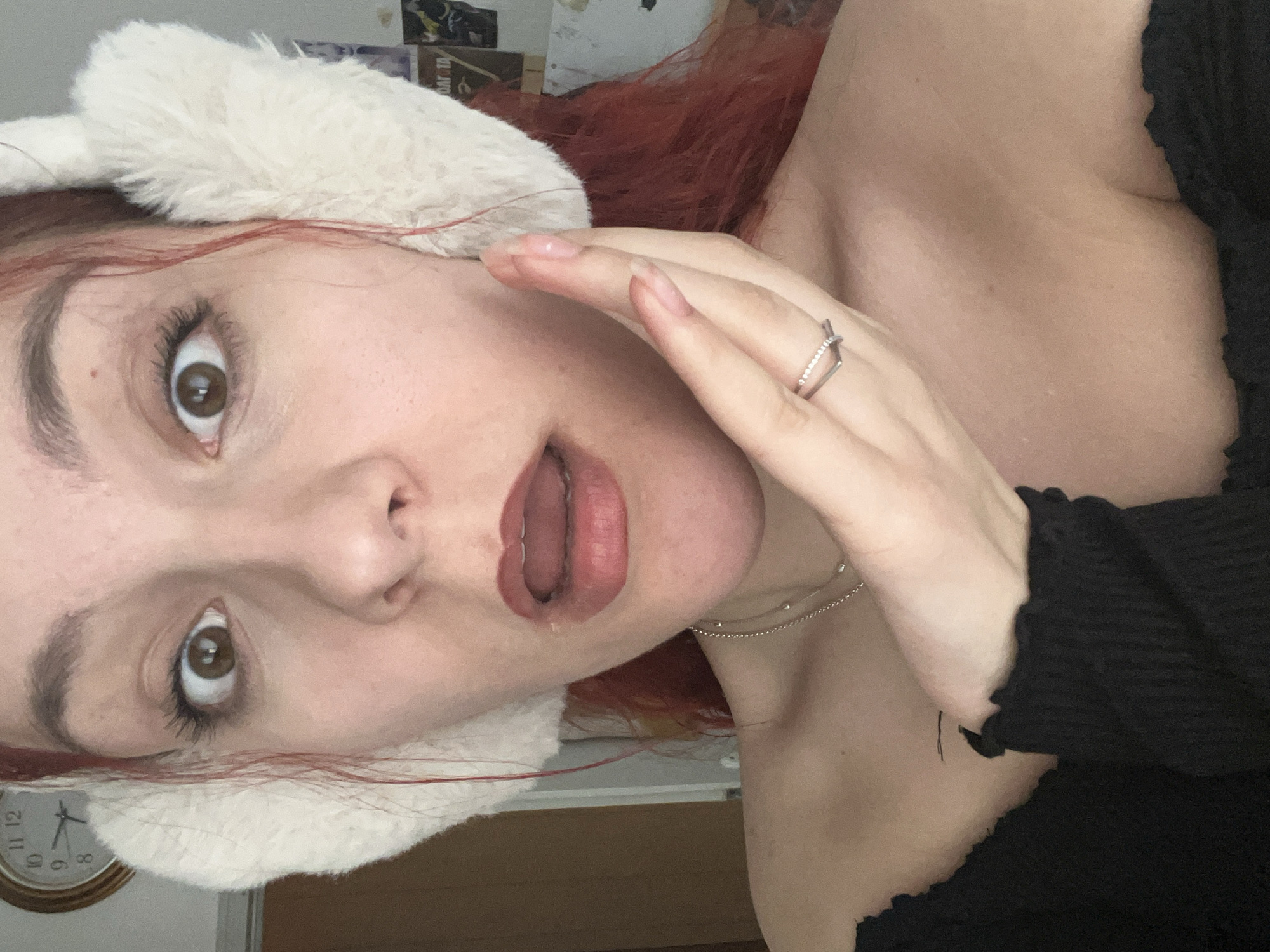 A close-up, angled portrait depicts a woman with fair skin, reddish-brown hair, and brown eyes. She is wearing a white furry head covering. Her right hand is raised near her face, displaying a silver ring on one of her fingers. She wears a delicate silver chain around her neck and a black top. A clock is visible in the background. The location is not discernible.