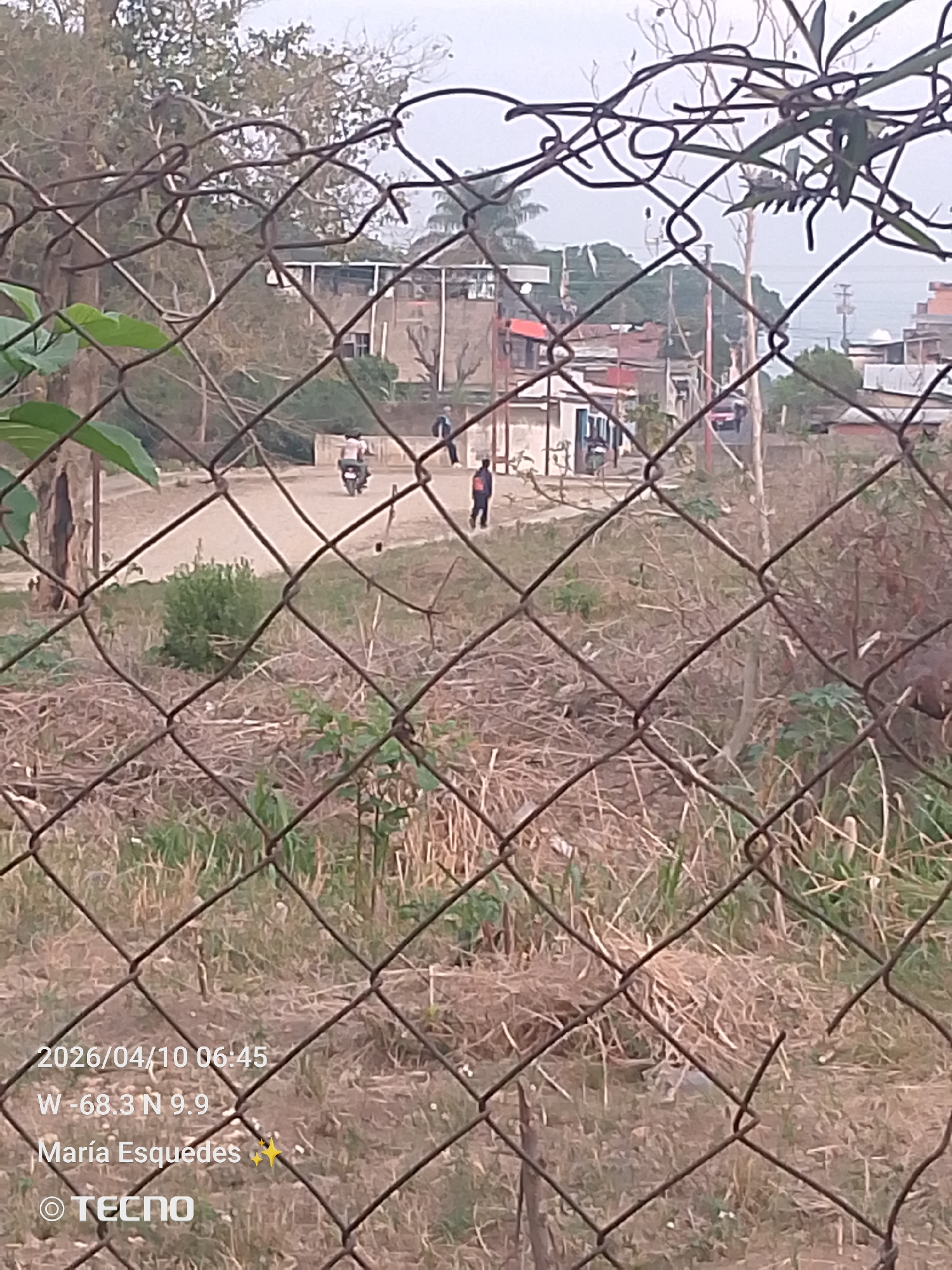 The image, dated 2026/04/10 at 06:45, taken at coordinates W -68.3 N 9.9, and attributed to María Esquedas, depicts a scene in Tinaquillo, Venezuela, viewed through a chain-link fence. In the midground, a dirt road intersects with other roads, on which a motorcyclist is riding. There are two individuals walking. Various buildings are visible, indicating a residential or semi-urban environment. The foreground contains dry vegetation and uncut grasses. The sky appears overcast.