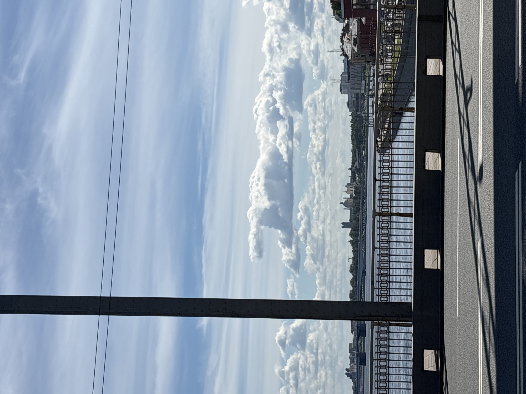 This is a vertical, low-angle shot looking out from a bridge in Saint Petersburg, Russia. The image captures a panoramic view of the city's skyline and the Neva River under a dynamic sky filled with dramatic, billowing clouds. The foreground is dominated by the dark metal structure of the bridge, with two parallel black cables stretching across the sky. To the right, the asphalt road of the bridge is visible, marked with white lines and casting long shadows from an unseen object.
Beyond the bridge railing, the Neva River flows, reflecting the sky and dotted with boats. Along the riverbanks, the cityscape of Saint Petersburg unfolds. Numerous buildings, including churches with domes and modern structures, are visible. Lush green trees line the riverbanks, creating a verdant contrast with the urban architecture. The weather appears pleasant, with a mix of bright sunshine and fluffy white clouds, suggesting a clear and possibly warm day. The time of day is likely midday, given the strong shadows. There is no visible text in the image. The overall impression is one of a grand urban landscape viewed from an elevated perspective on a beautiful day.