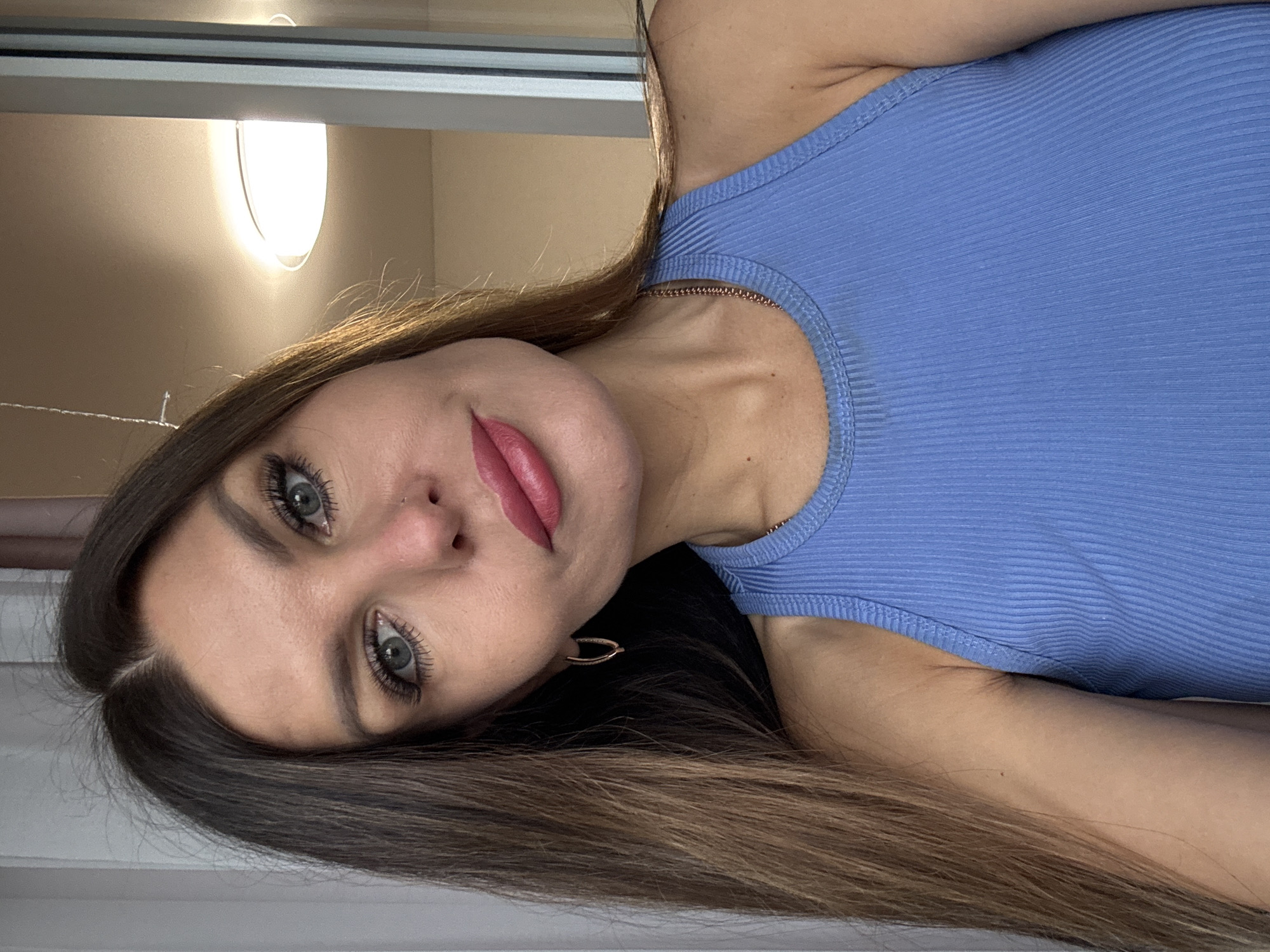 A woman with light olive skin and brown hair is facing the camera. She has green eyes and full pink lips, with a piercing on the left side of her nose. She is wearing a blue ribbed tank top and gold earrings. The background shows a white wall and a round light fixture near a window. The location is unknown.