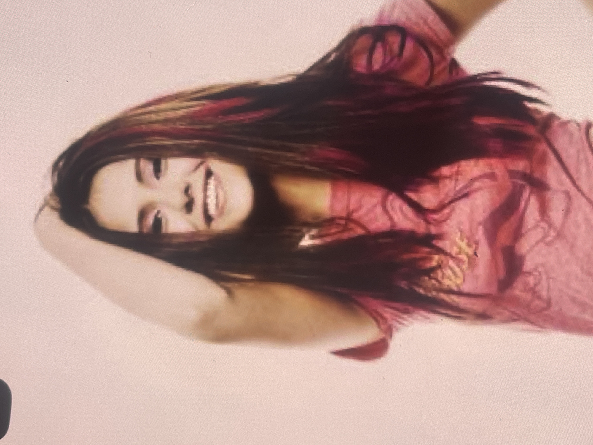 This medium-shot image, rotated 90 degrees clockwise, captures a young person, likely female, posing with a joyful expression against a plain, light off-white or pale pink background. The setting is indoors, possibly a studio in Poltava, Ukraine.
The subject has long, dark hair with prominent red streaks and some lighter, possibly blonde or light brown, highlights. They are smiling broadly, showing their teeth, indicating happiness and playfulness. One arm is bent and raised, with the hand positioned near their head, contributing to a relaxed and candid pose. Their hair cascades around their head and shoulders.
The individual is wearing a pink t-shirt that features a blurry yellow or gold graphic design on the front, though no specific text is discernible. The image itself appears to be a photograph of a screen or a digital capture, showing some pixelation and a slightly warm, faded quality. The lighting is soft and consistent, emphasizing the subject's features and bright smile.