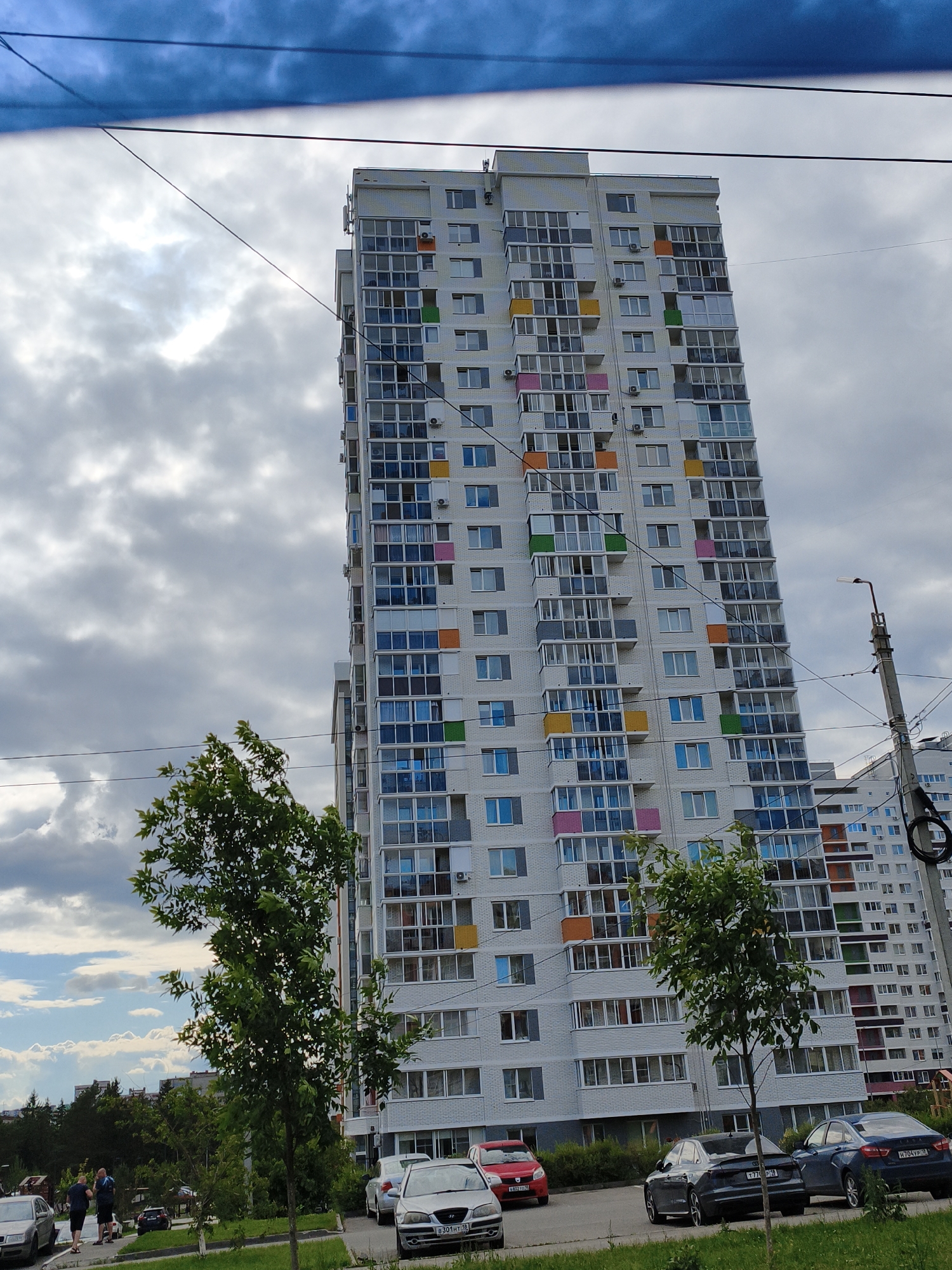 The image captures a view of a tall, modern apartment building in Pervomaysky, Russia, under a cloudy sky. The building is white with numerous windows and balconies, many of which feature colorful accents in orange, yellow, green, and pink. Power lines crisscross the foreground. To the left of the apartment building, a smaller building is visible, and further to the left, trees and a few cars are present. In the foreground, parked cars are lined up along a street, and two men are standing and walking near them. The overall scene suggests an urban residential area.