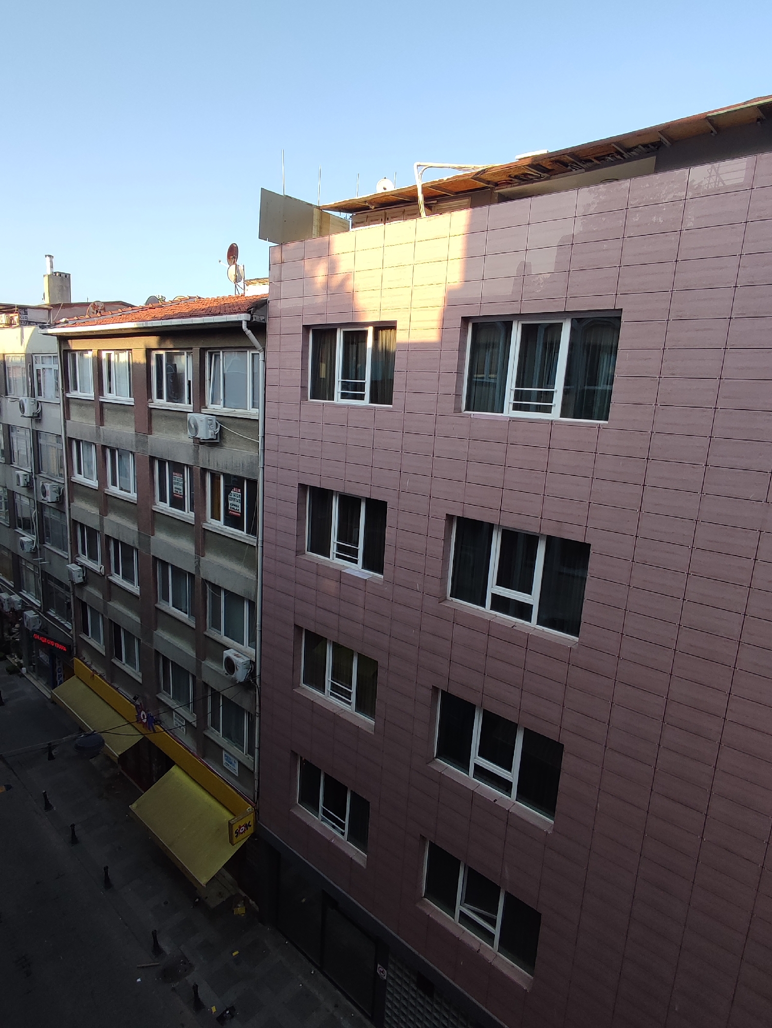 The image captures a street view in Kadıköy, Turkey, showing multiple buildings and a street below. On the right, a modern building with a pink tiled facade dominates the frame, featuring several rows of white-framed windows. To the left of this building, an older, grey concrete building is visible, also with multiple floors of windows and air conditioning units mounted on its exterior. The ground level of the grey building features shops with yellow awnings, one of which has a sign that appears to say "SOK." The street below is paved and has bollards lining its edge. The sky above is clear and blue, indicating daylight. The overall impression is of an urban street scene with a mix of architectural styles.