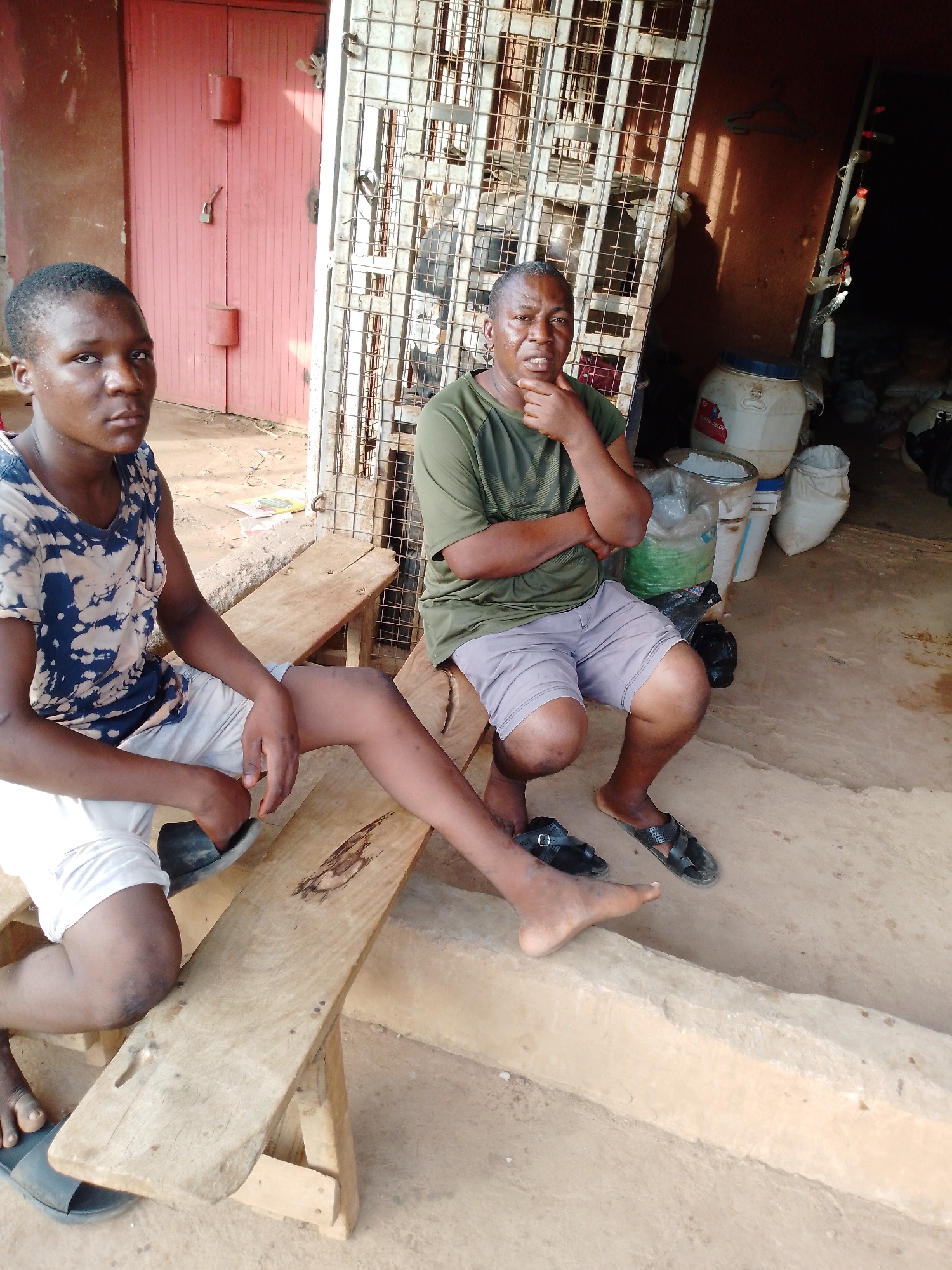Two males, an adult and a younger person, are seated on simple wooden benches in an outdoor or semi-outdoor setting during daylight hours.
On the left, the younger male with short dark hair is wearing a blue and white tie-dye short-sleeved shirt and white shorts. He has one black slide sandal on his right foot. He looks directly towards the camera with a neutral expression.
On the right, the adult male with short dark hair is wearing an olive green t-shirt and light gray shorts, with black sandals on his feet. He has his right hand resting under his chin, left arm crossed, and a contemplative expression as he looks slightly to the right of the frame.
Behind the individuals, to the left, is a closed red double door with visible locks and latches. To the right, a metal grate or gate provides a partial view into a darker area. This area contains several white buckets, plastic sacks, and a large white barrel, some appearing to hold white granular substances. Partial text "CHOLCALCI" and "D Cal" is visible on the white barrel.
The ground appears to be unpaved, a mix of dirt and possibly concrete. The lighting suggests a bright, sunny day, with visible shadows. The scene has a functional and informal ambiance, possibly depicting a waiting area or a storefront entrance.