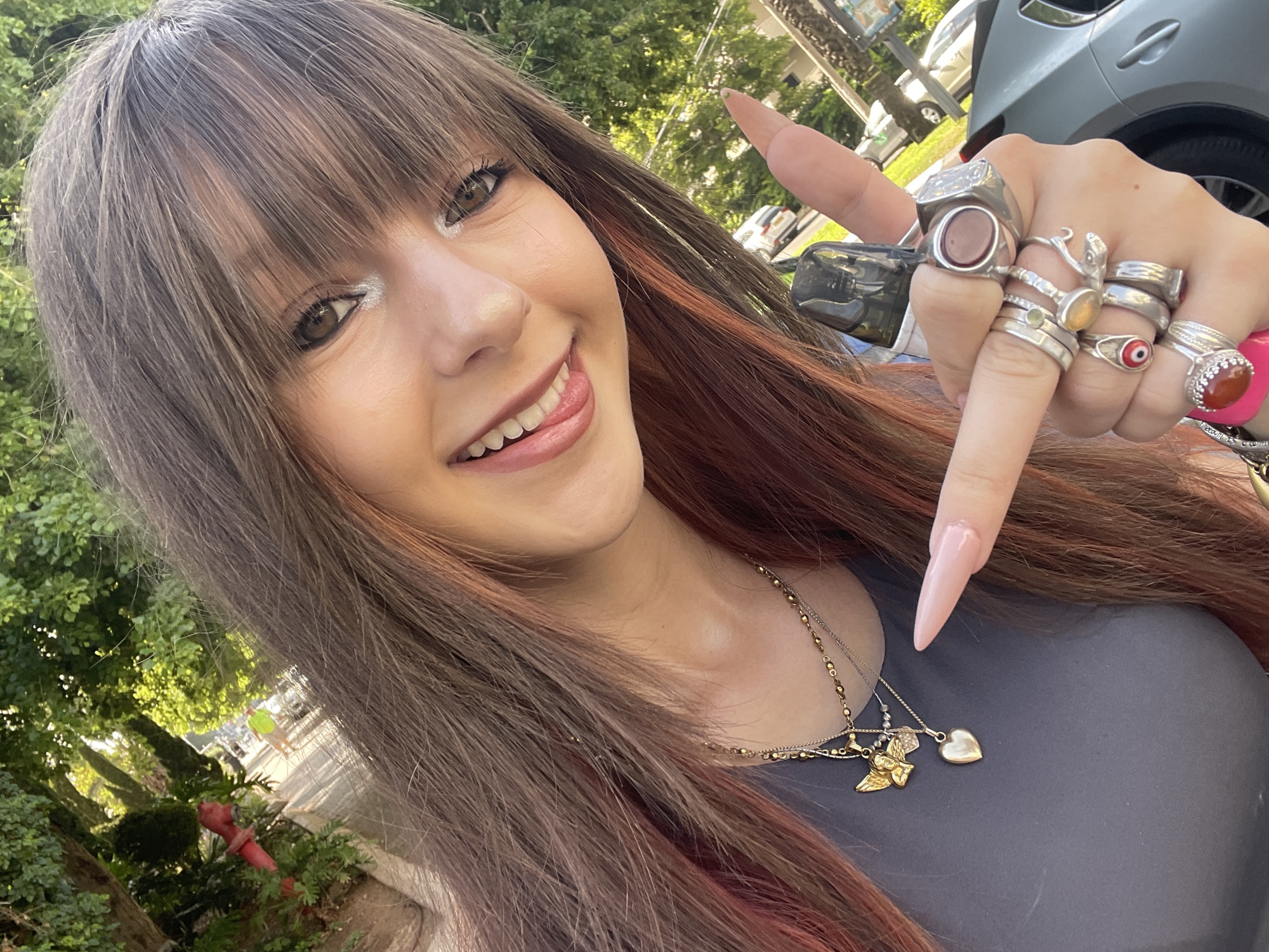 A young woman is captured in a close-up selfie outdoors. She has long, brown hair with streaks of pink, and her bangs partially obscure her eyes. She is wearing a gray t-shirt and multiple necklaces, including one with an angel and heart pendant. She has several rings on her fingers, including a large ring with a reddish stone on her right index finger. Her nails are long and painted a light pink. She is smiling and sticking her tongue out slightly, with her right index finger pointing towards the viewer. The background suggests an outdoor setting with trees and a street, with cars visible in the distance. The lighting indicates it is daytime. This scene does not depict a specific location in Raanana, Israel.