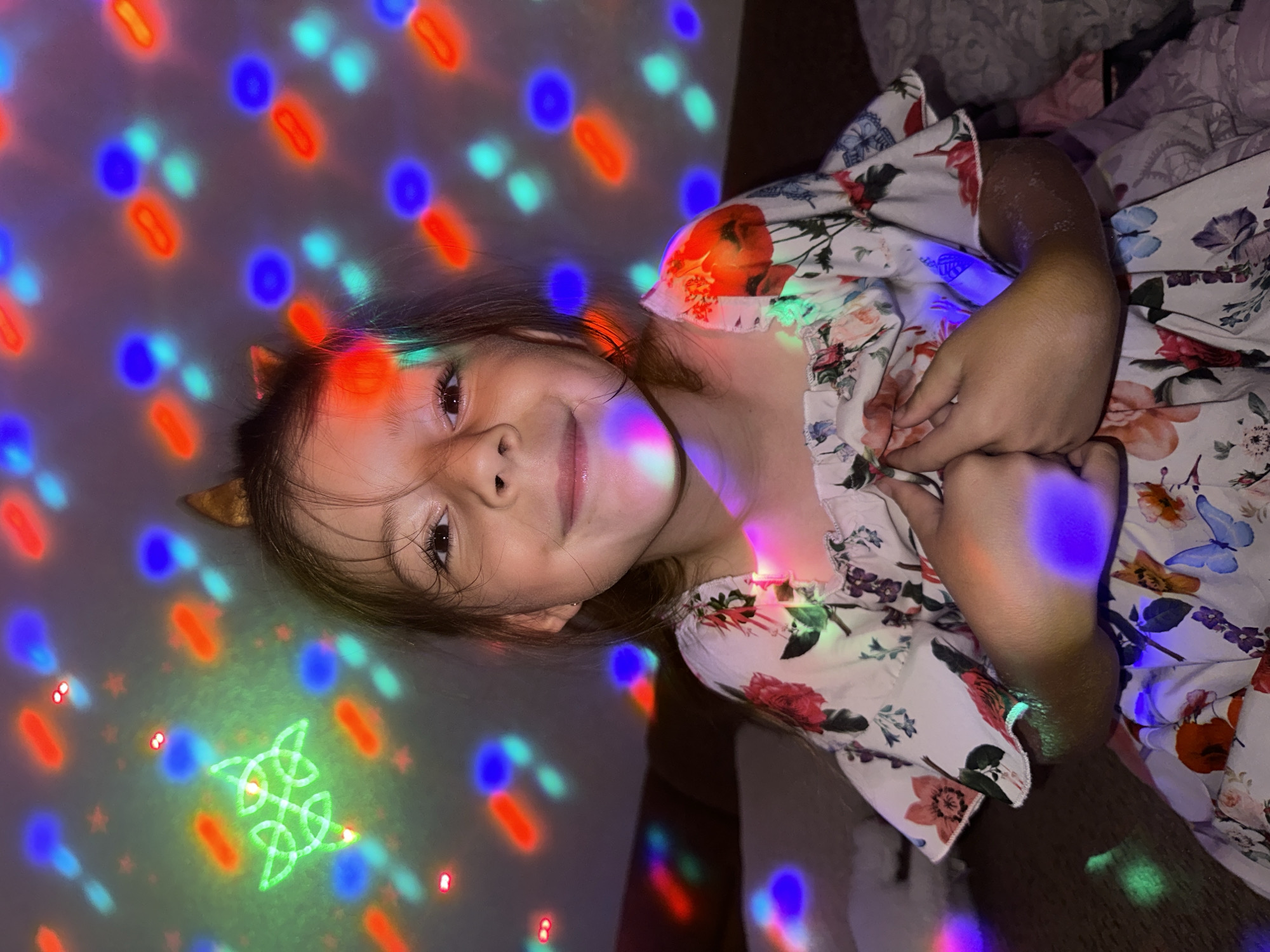 A young child, appearing to be between four and eight years old, is seated indoors, smiling directly at the camera. The child has light brown hair and is wearing a white, off-the-shoulder dress with a multi-colored floral pattern featuring red roses, blue butterflies, and green foliage. A golden-brown, ear-shaped headpiece is visible on top of the child's head. The child's hands are clasped together in front of her chest. The background, an interior wall, is dark, illuminated by numerous projected colored lights. These projections include out-of-focus red oval shapes, blue circular shapes, and teal oval shapes, along with smaller red dots. In the upper left quadrant of the image, a distinct green laser projection forms an intricate, symmetrical knot design. Various colored lights from these projections reflect onto the child's face, hair, arms, and dress. The scene takes place in an indoor location in the city of Esenyurt, Turkey.
