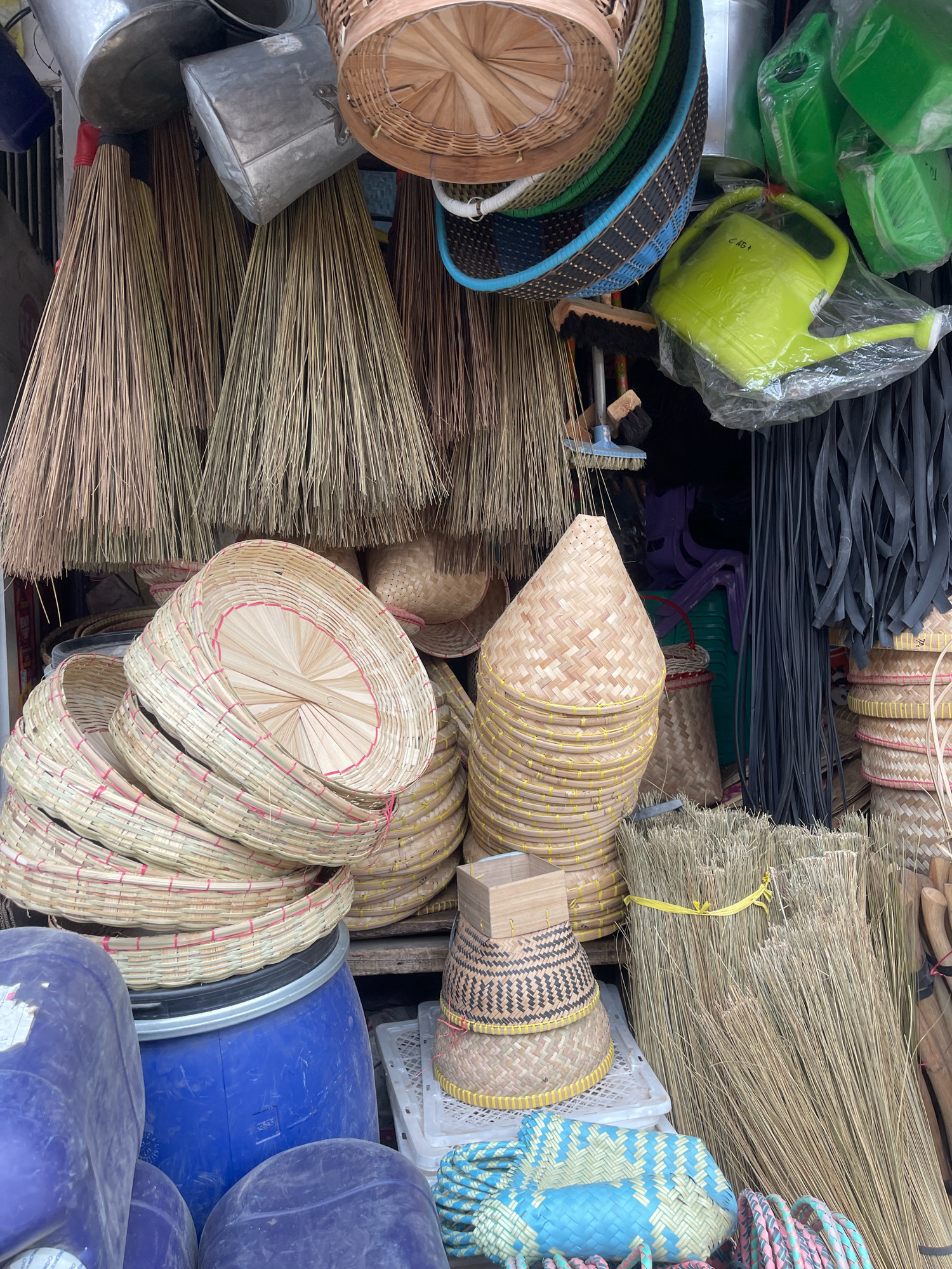 This is a medium-shot, eye-level, natural light, exterior scene of a street market stall in Sukabumi, Indonesia, likely selling household and gardening items. The foreground is dominated by various woven bamboo and rattan baskets, including several large, shallow circular ones stacked on top of each other and a tall, conical woven container with yellow banding. To the left, large, blue plastic barrels are visible, along with two large, dark purple plastic containers. In the center-right, a large bundle of dried grass, presumably for brooms, is tied with a yellow ribbon. Behind these items, a collection of brooms with wooden handles and straw bristles hangs from above. Further back, a lime green watering can is visible, still in its plastic packaging, along with several other green plastic items, some of which appear to be smaller watering cans or containers. The background shows more stacked woven baskets of various sizes and colors, as well as black rubber strips hanging vertically. The overall impression is of a well-stocked stall offering a variety of rustic and practical goods.