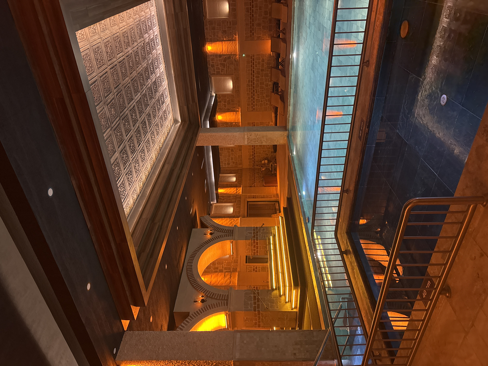 The image depicts a subterranean space in Ürgüp, Turkey. Visible architectural elements include stone walls with arched openings, a decorative ceiling with patterned tiles, and what appears to be a shallow pool or reflective surface with tiled edges. A metal railing and staircase are also visible. The space is illuminated with warm, artificial lighting, creating a yellow-orange ambiance. The scene is devoid of people.