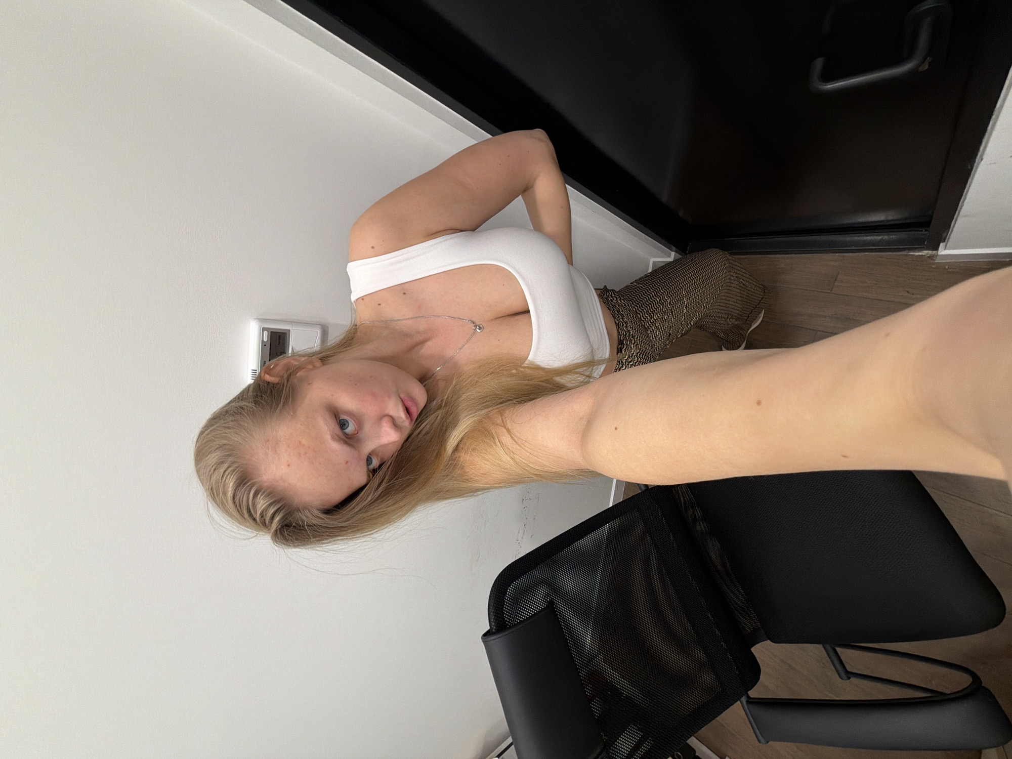 A selfie-style photograph, rotated 90 degrees clockwise, captures a young woman with long blonde hair and blue eyes. She is wearing a form-fitting white tank top and brown and black patterned trousers, along with a delicate silver necklace featuring a small charm. Her gaze is directed straight at the camera, and her expression is neutral.
The background indicates an indoor setting. To her left, a plain white wall features a white wall-mounted control panel, possibly a thermostat or vent. Behind her, a dark, likely black, door with a metallic handle is visible, and the floor beneath appears to be made of wood. Part of a black office chair with a mesh back is positioned in the foreground and to her right. The overall impression is a modern interior space, potentially within an apartment or office in Izhevsk, Russia. No discernible text is visible in the image.