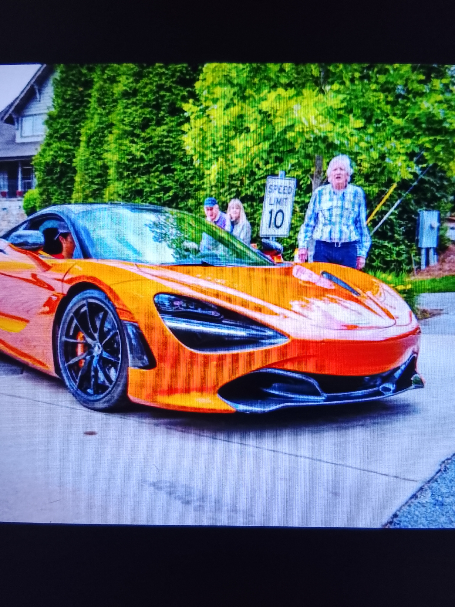 In an outdoor setting, possibly a residential street, a bright orange McLaren sports car is parked. The car's sleek design and low profile are prominent. Behind the car, a white-haired elderly woman wearing a blue and white plaid shirt and dark pants stands beside a speed limit sign that reads "SPEED LIMIT 10". To her left, a man in a blue cap and a woman with blonde hair are partially visible, seemingly interacting with the scene. In the background, a large house with a pitched roof and dense, green evergreen trees line the street. The overall atmosphere suggests a casual gathering or a moment of admiration for the high-performance vehicle.