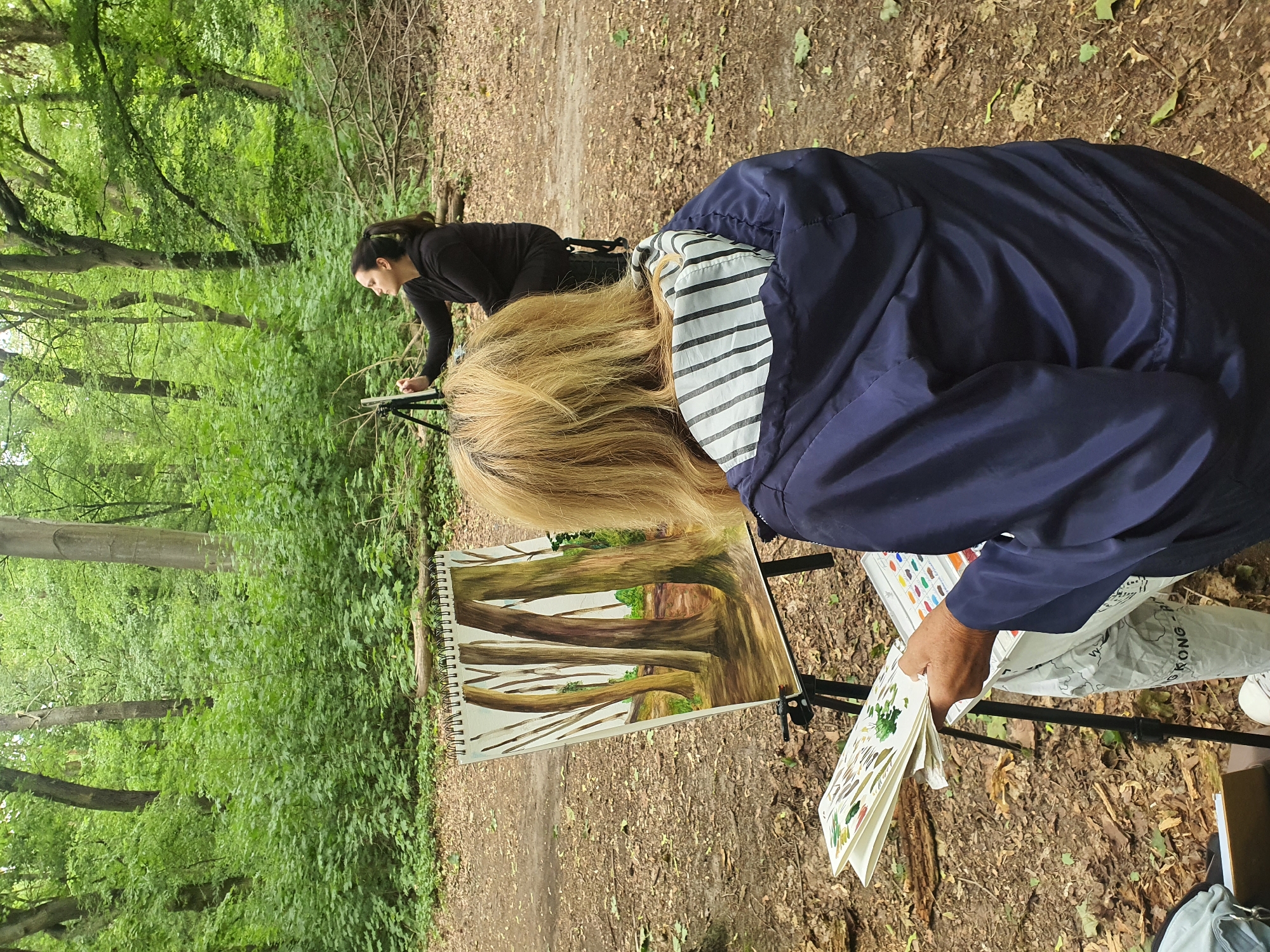 Two individuals are engaged in painting outdoors in a wooded area, likely a park or forest, in Falkensee, Germany. In the foreground, a person with blonde hair is seated and looking at a sketchbook and a painting on an easel. The painting on the easel depicts several tree trunks with a background of greenery. The person is wearing a dark blue hooded jacket with a striped lining around the hood. In the background, another person is seated on a stool, facing away from the viewer, and appears to be painting on a smaller easel. The environment is characterized by numerous trees and fallen leaves on the ground, suggesting an autumn or spring setting. The light suggests it is daytime.
