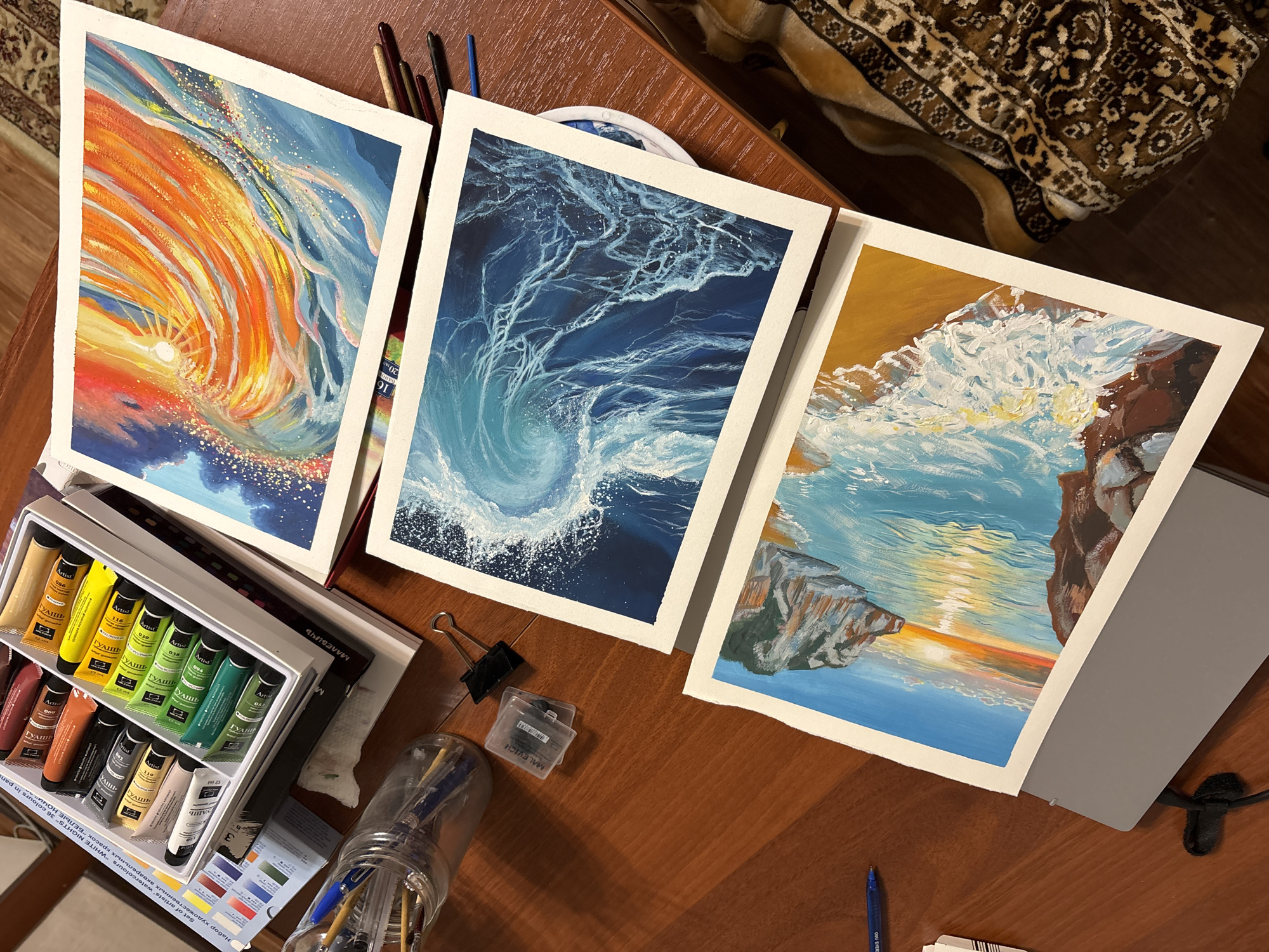 The image depicts a wooden table surface displaying three watercolor paintings. A set of watercolor paints, labeled "Artist" and "White Nights," is positioned next to the paintings. The paints are of various colors. A jar holding paint brushes and additional painting tools are also visible. The background includes a carpet. The location in the city of Izhevsk, Russia is not explicitly shown, and therefore cannot be identified in the image.