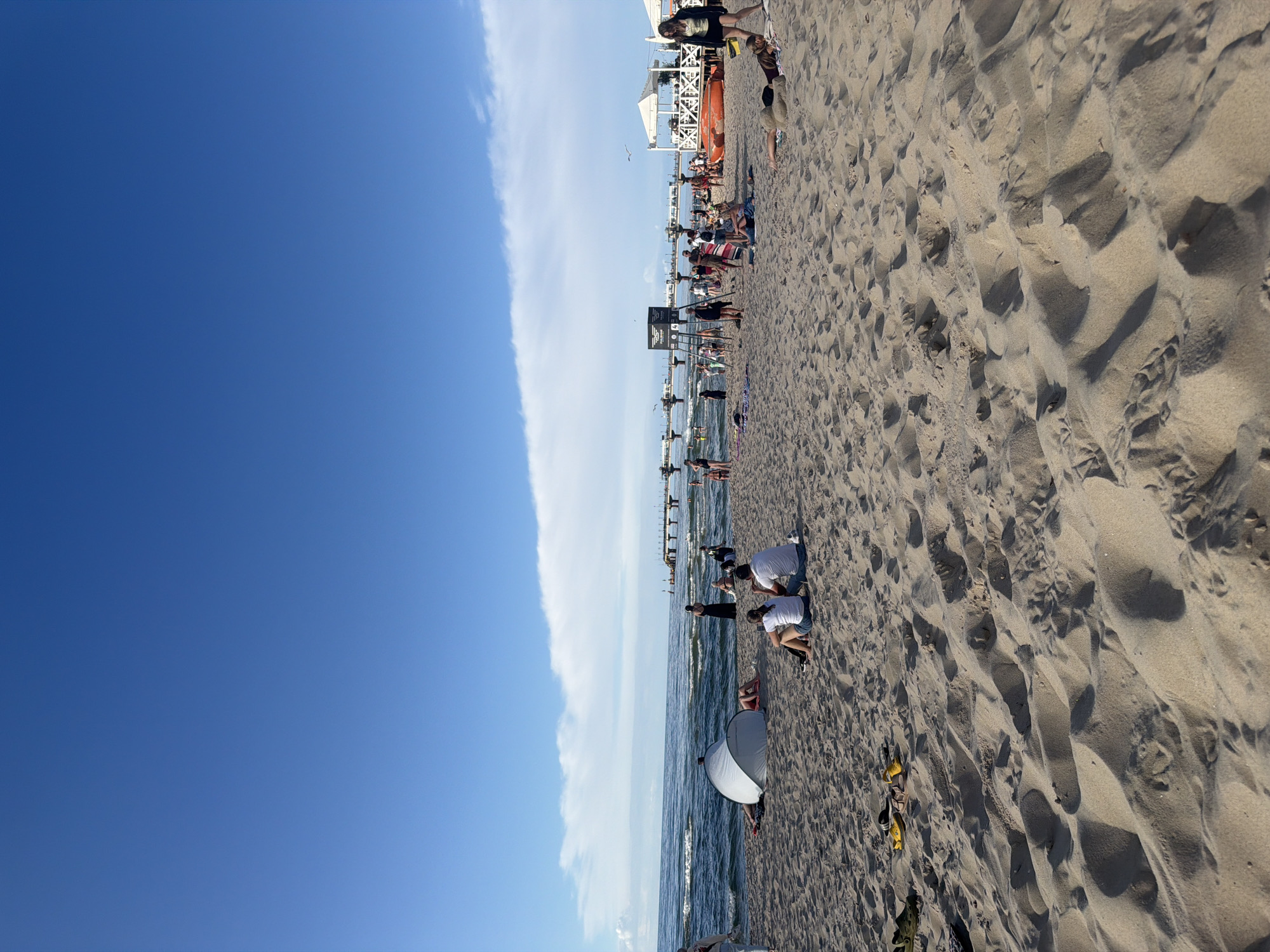 This image depicts a sunny day at the beach in Międzyzdroje, Poland, with the Baltic Sea and a pier visible in the background. The sandy beach is populated with numerous people engaged in various beach activities. In the foreground, a white tent is set up on the sand, and several individuals are seated on the beach, facing the sea. Further back, a crowd of people are scattered across the beach, some near white structures that appear to be umbrellas or gazebos. A long pier extends into the sea, with people walking on it. The sky is a clear blue, with a few white clouds. The overall scene captures a lively atmosphere at a popular seaside resort.