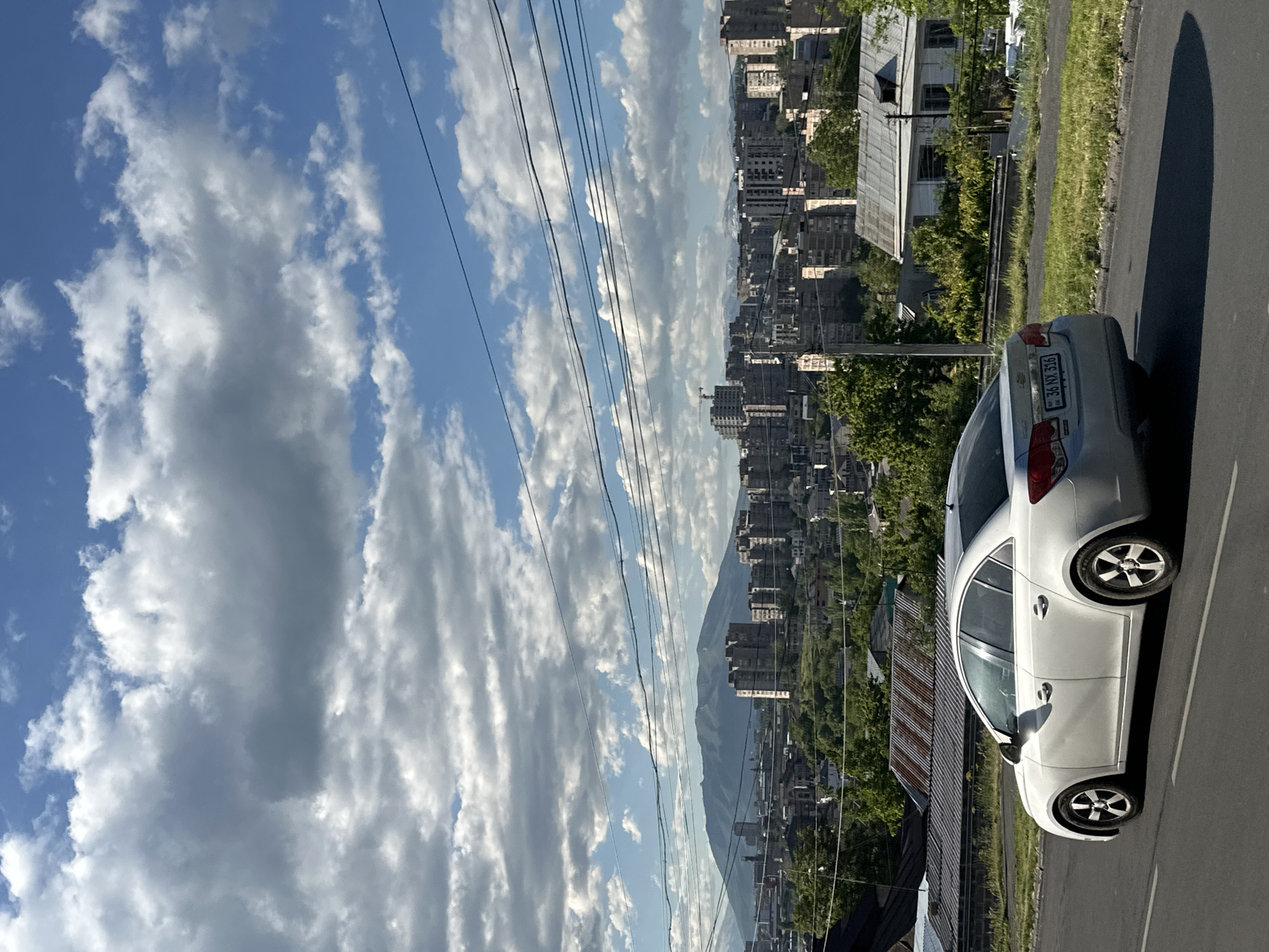 The image depicts a cityscape, seemingly taken from an elevated position in Yerevan, Armenia, as evidenced by the license plate of the white sedan (license plate 36 NX 316). The sky is filled with bright white clouds against a blue backdrop, with power lines running across the field of view. Below, a densely populated urban area unfolds, featuring a mix of low-rise and high-rise buildings and the suggestion of a mountain range in the distance. In the foreground, a white sedan is parked along a road; its license plate is visible. Trees and other buildings also are present in the foreground.