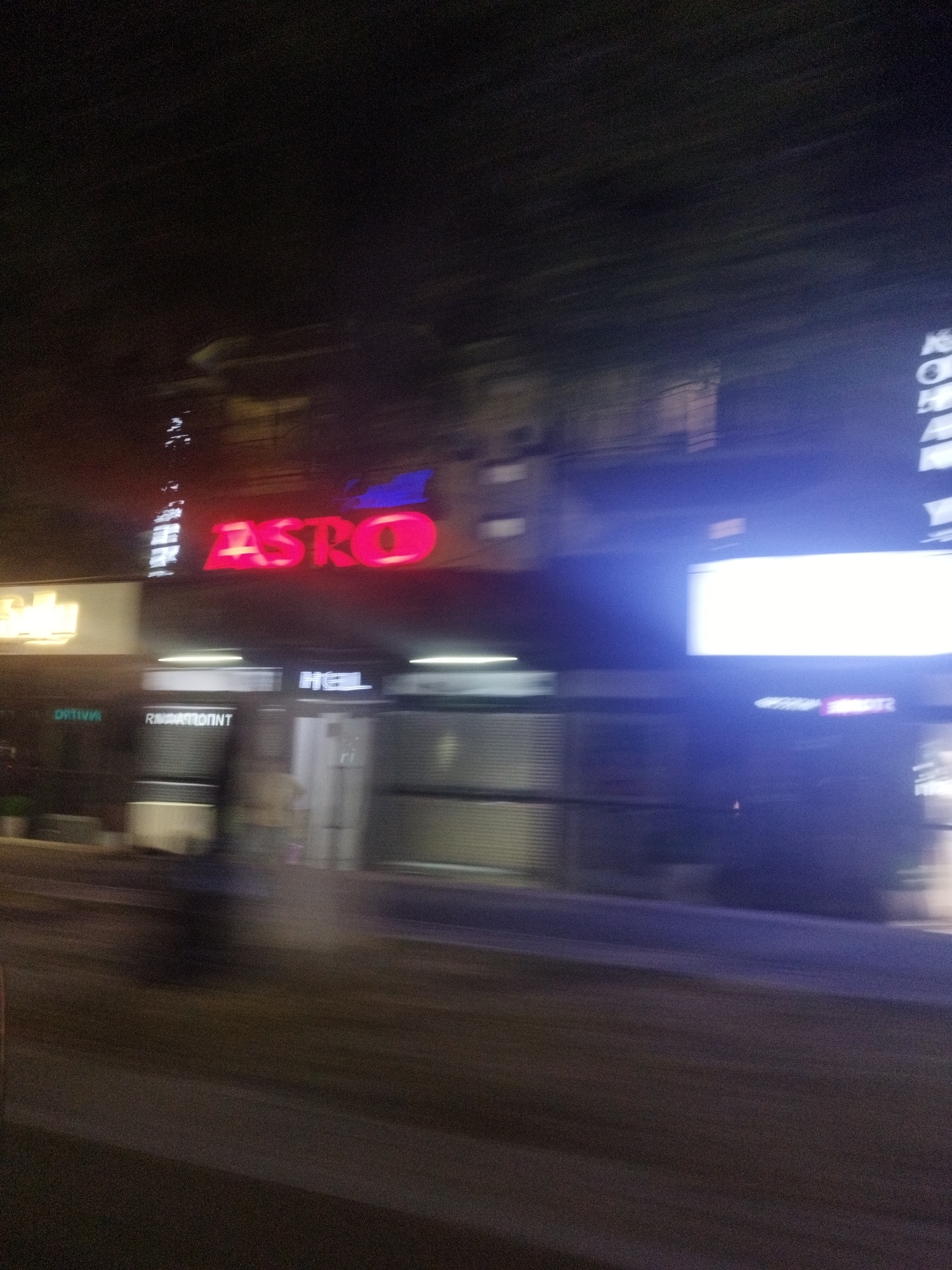 The image is a blurry, night-time photograph of a street scene in Shymkent, Kazakhstan. A prominent red neon sign displaying the word "ASTRO" is visible. Below this sign, another sign appears to read "OPTIVA" and another reads "RESTAURANT". Further to the right, a sign with white letters reads "HOTEL". There is a large, bright white illuminated display screen to the right of the "HOTEL" sign. The street in front of the buildings appears to be paved. The overall impression is of a commercial street at night with illuminated signage.