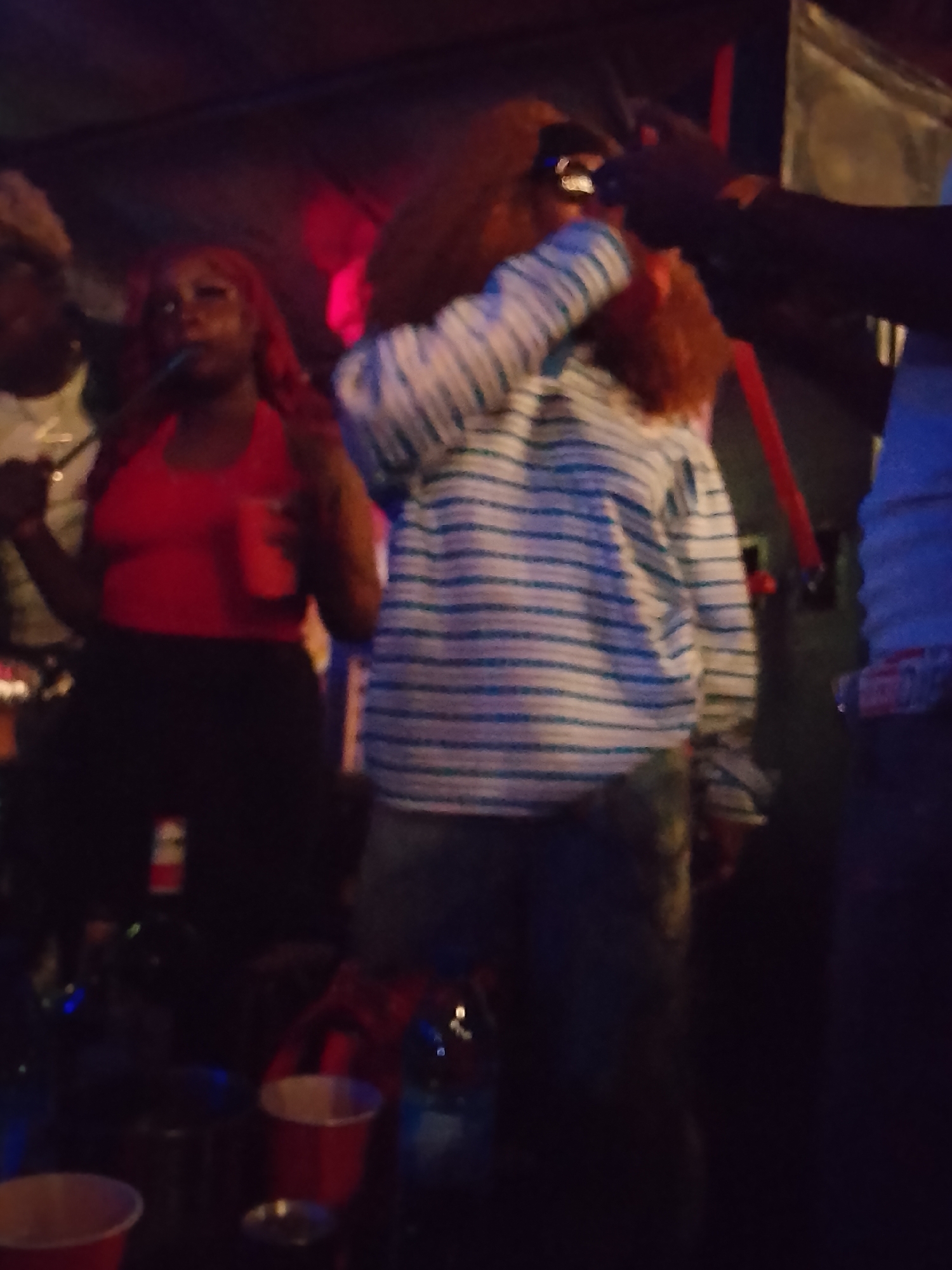 The image appears to be taken at a dimly lit gathering, possibly a party, in what appears to be an indoor space, given the presence of indoor lighting. There are multiple individuals present. A woman in a red top and dark pants is holding a red cup. Another individual wearing a blue and white striped shirt is shown with their head turned as they appear to be drinking from a bottle. There are red plastic cups and bottles on a surface, possibly a table, in the foreground. The overall scene suggests a social gathering with possible alcohol consumption. No further information regarding the specific location in Narayi, Nigeria, can be determined.