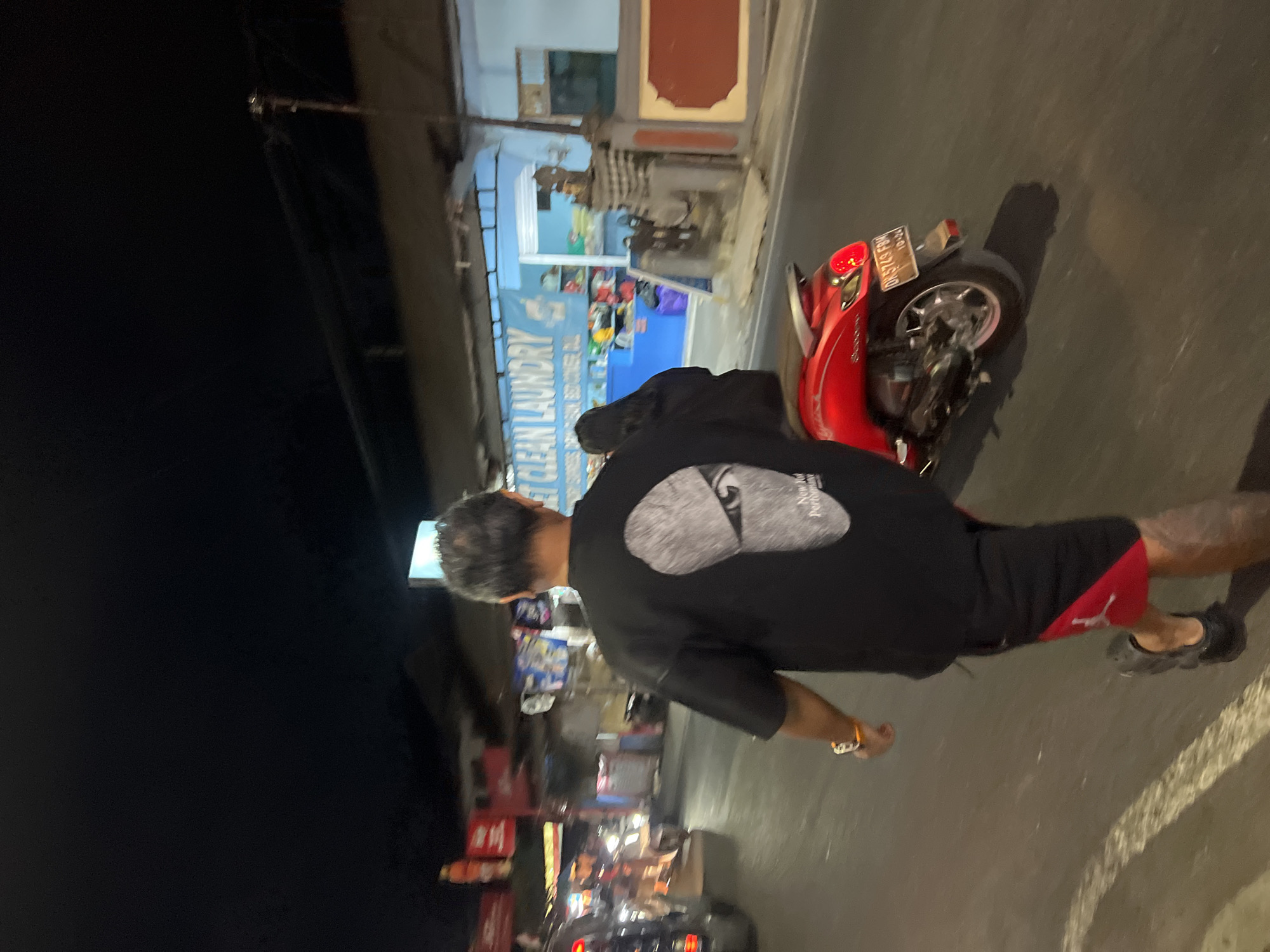 In the city of Denpasar, Indonesia, at night, a man wearing a black t-shirt with a graphic design on the back and red and black shorts walks on a street. He is walking away from the camera and is near a red scooter. To his left, a woman with long dark hair is visible. In the background, a building with a blue sign that says "CLEAN LAUNDRY" is present, along with other street lights and vehicles. The street appears to be paved.