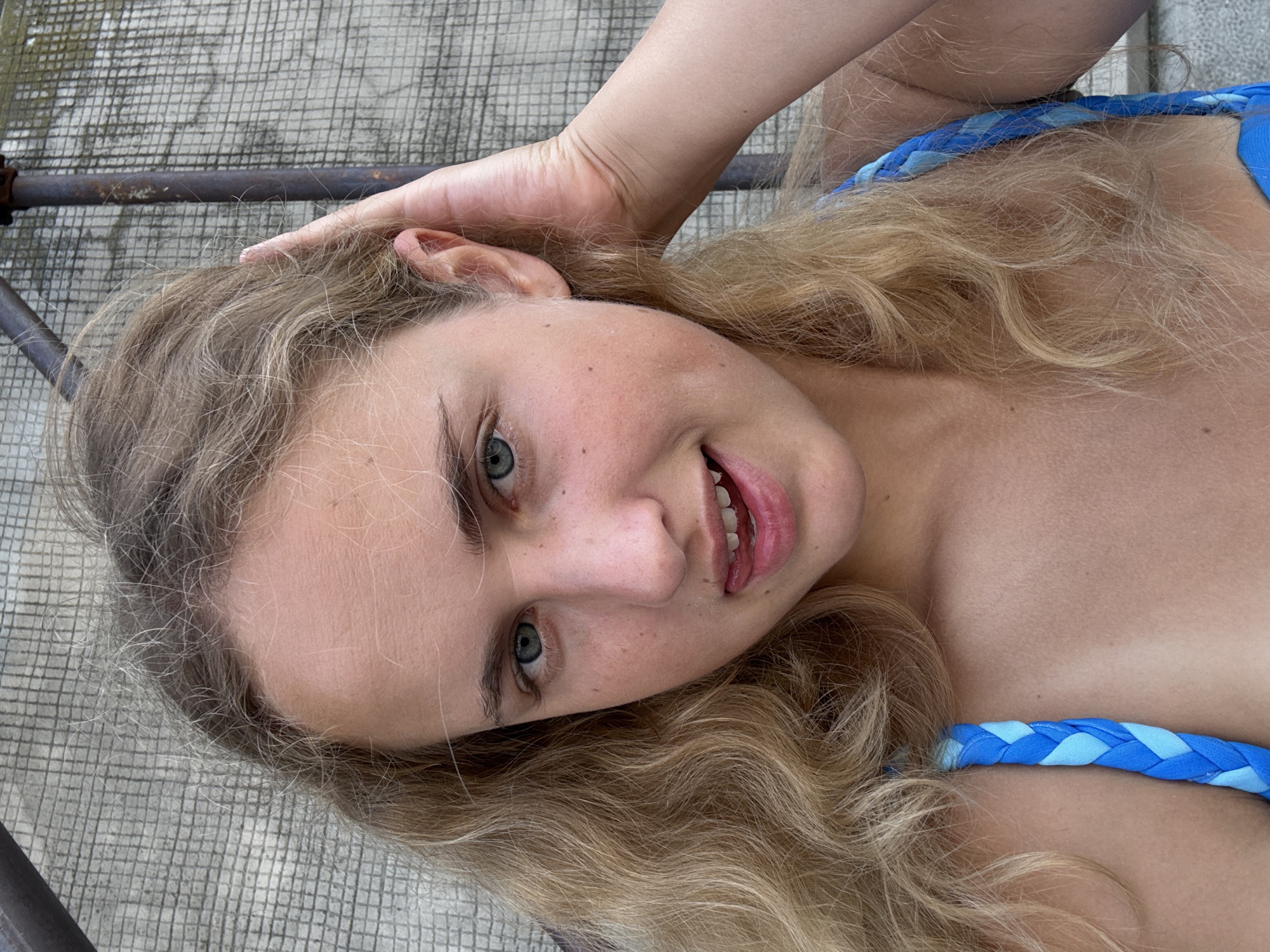A young woman with blonde hair and blue eyes is pictured outdoors against a grey, grid-patterned wall. She is looking at the camera and her right hand is raised, touching her hair near her ear. She is wearing a blue braided bikini top. Her mouth is slightly open, and her tongue is visible, touching her lower lip. The image is a close-up, focusing on her face and upper body. The lighting suggests it is daytime. The location is not specified in the image or provided information.