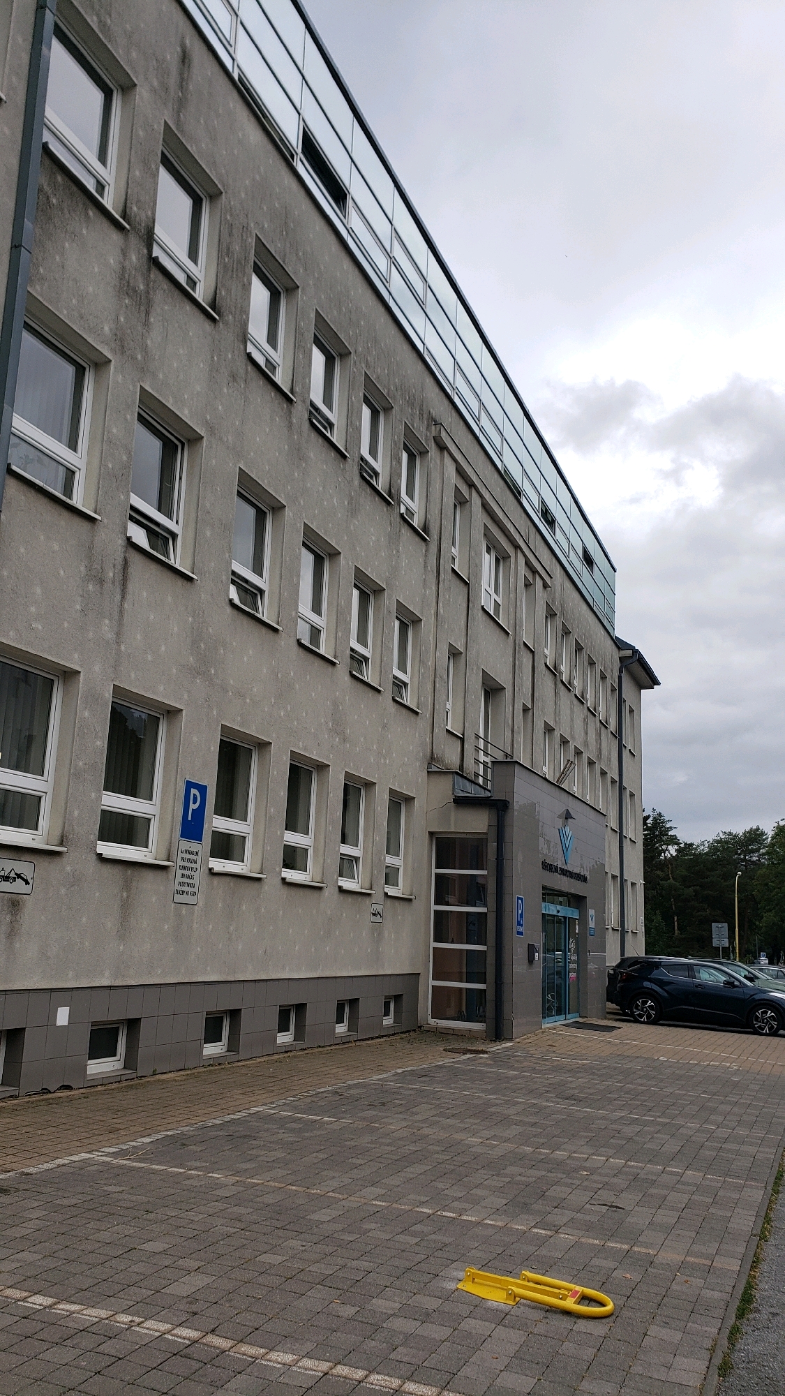 The image shows the exterior of a modern building with a grey facade and a glass-enclosed upper level. The building has numerous windows and an entrance with glass doors. A dark-colored car is parked in front of the building. The foreground consists of a paved parking lot. The sky is overcast. The building is identified as the Regional Office of the Slovak Land Survey and Cadastre Authority in Prešov, Slovakia, located at Jesenná 13. There are no people visible in the image.