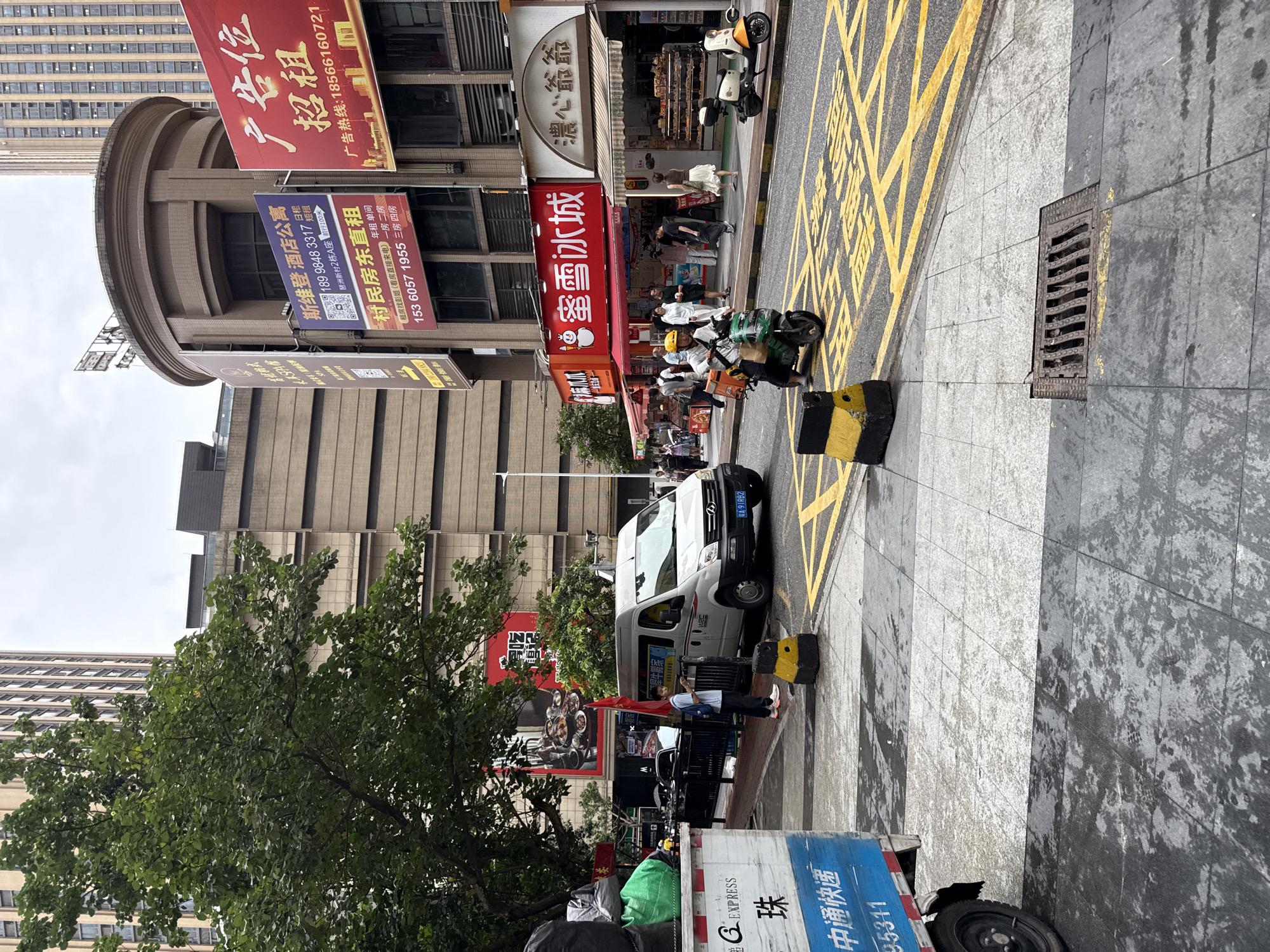 This image depicts a street scene in Haizhu District, China. A white van is parked on the right side of the road, with a person standing near its rear door. A person on a green scooter with a yellow delivery box is moving through the intersection. To the left, a large blue and white truck is partially visible. A variety of businesses line the street, including one with a prominent red sign featuring a cartoon snowman and Chinese characters that read "Xue Bing Cheng" (Ice City). Another shop displays baked goods. Buildings of varying heights, some with signage, dominate the background. The street appears to be paved with gray tiles and features yellow traffic markings. The sky is overcast.