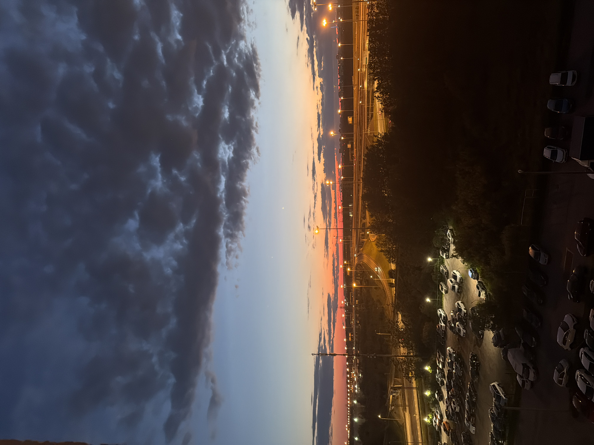 The image shows a parking lot filled with cars at dusk. The sky is a deep blue with streaks of orange and red from the setting sun. Streetlights illuminate the parking lot, casting long shadows. In the background, a highway overpass is visible, with cars driving on it and streetlights lining the road. The overall scene suggests a suburban or urban environment.
