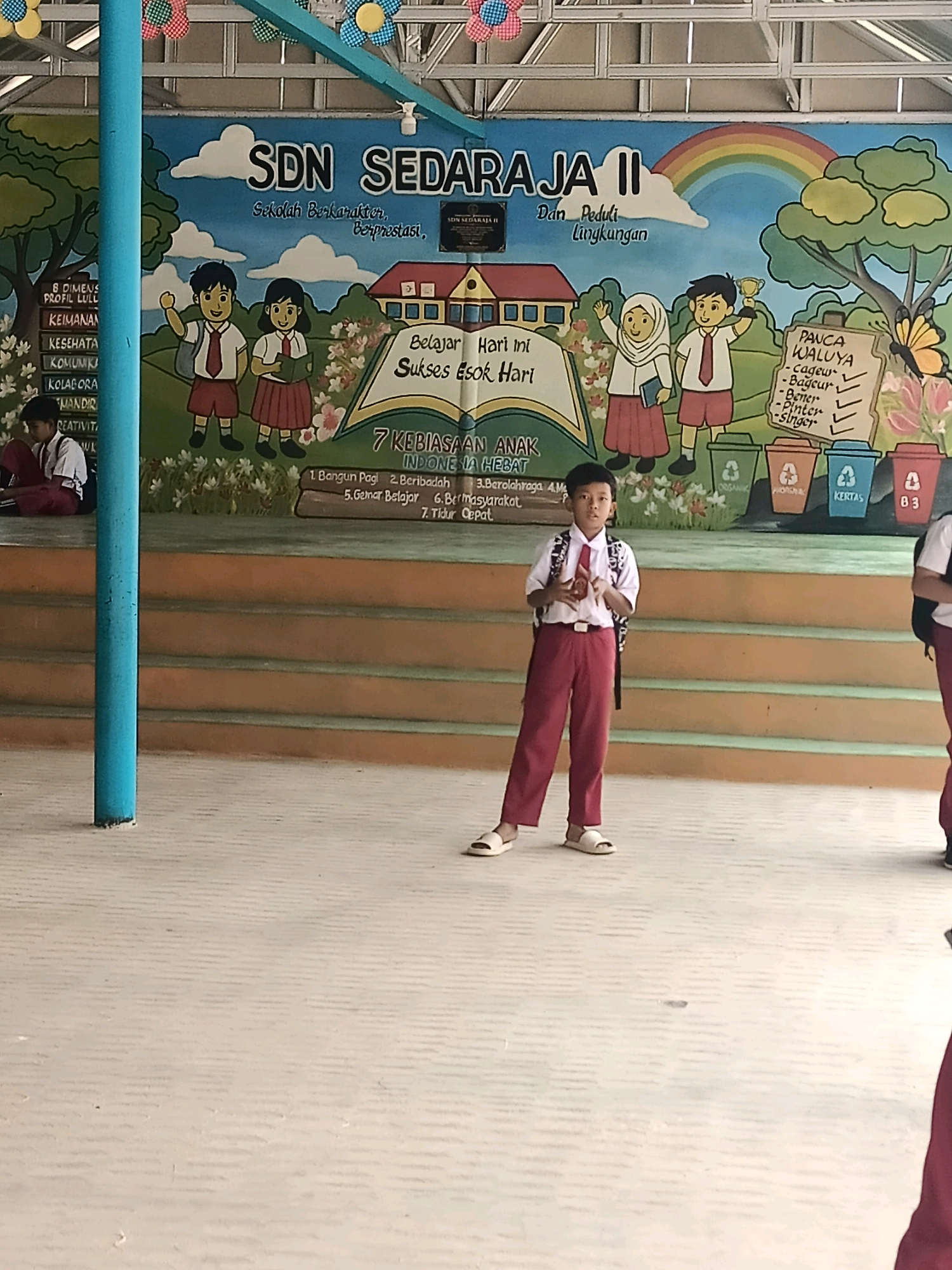 The image depicts the facade of SDN Sedaraja II, an elementary school in Sedaraja, Indonesia. A vibrant mural adorns the wall, featuring cartoon characters, educational slogans, and a rainbow. In the foreground, two male students in school uniforms stand on a paved area. The student on the left is wearing a white shirt, red tie, and red pants, with a backpack slung over his shoulder, and is holding something in his hands. The student on the right is partially visible, also in a red and white uniform. The school building is visible in the background, with tiered seating in front of the mural. The environment appears to be an outdoor schoolyard.