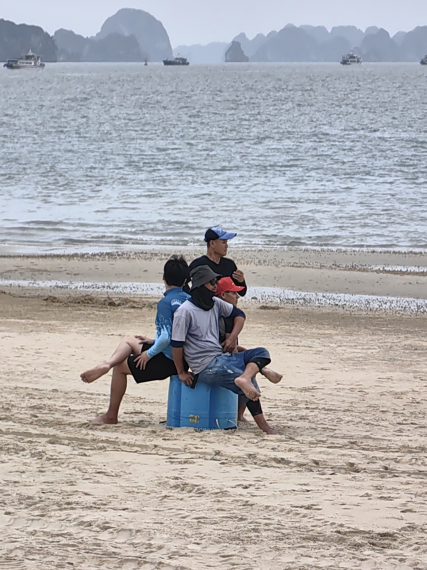 This media file depicts a coastal scene in Bai Chay Ward, Vietnam, likely within the broader Ha Long Bay area given the distinctive limestone karsts. The main subjects are four males gathered on a sandy beach.
In the midground, three of the men are resting around a blue plastic barrel. One man, wearing a blue long-sleeved shirt and black shorts, is seated on the sand, leaning against the barrel with his back partially to the viewer. Another man, in a grey t-shirt, blue jeans, and a dark face covering, is sitting directly on the blue barrel, facing towards the right. His bare feet are visible, crossed at the ankles. A third man, wearing a red cap and a dark shirt, is seated partially behind the man on the barrel, also facing right. A fourth man, dressed in a black t-shirt and a blue baseball cap, stands or leans near the group, looking towards the right side of the frame. The men appear to be resting or engaged in conversation.
The immediate foreground shows light-colored sand with minor undulations and footprints. Beyond the people, the calm, grey-green sea extends to the horizon, dotted with several boats. In the far background, numerous large, rugged limestone islands and mountains rise prominently from the water, a signature feature of the Ha Long Bay region. The sky is uniformly overcast, casting a soft, diffused light over the scene and contributing to a hazy atmospheric effect. The time of day is not precisely clear but suggests either early morning or late afternoon due to the subdued lighting. The blue barrel has some faint yellow markings or text, possibly "5G" or a similar short inscription, but it is largely illegible. The overall impression is one of quiet repose in a picturesque natural setting.