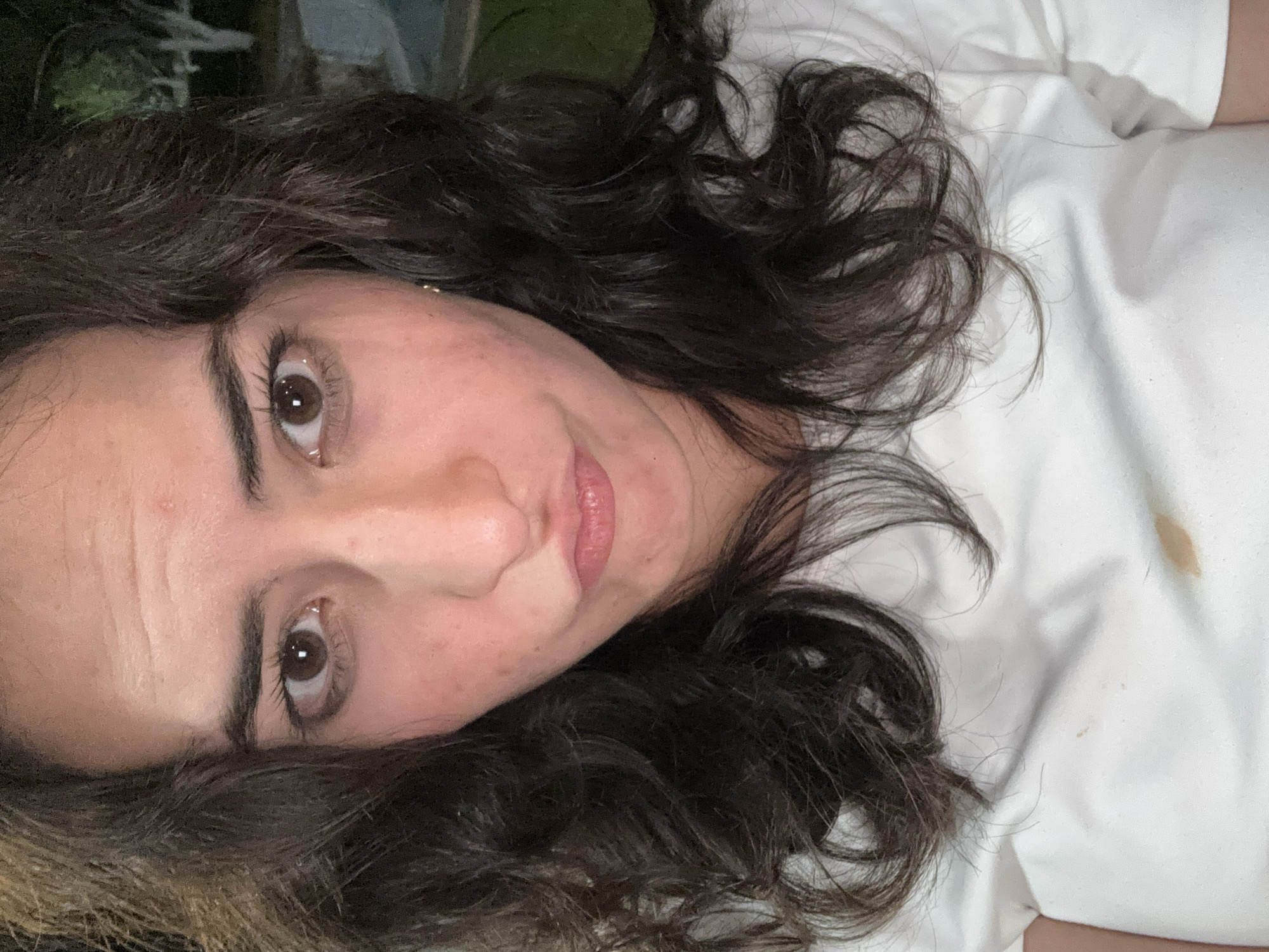 A woman with brown eyes and dark, curly hair lies on a white surface, possibly a bed. Her face is angled towards the camera. A stain is visible on the white surface near her right shoulder. The location is unknown and cannot be determined to be Puebla, Mexico, from the image alone.