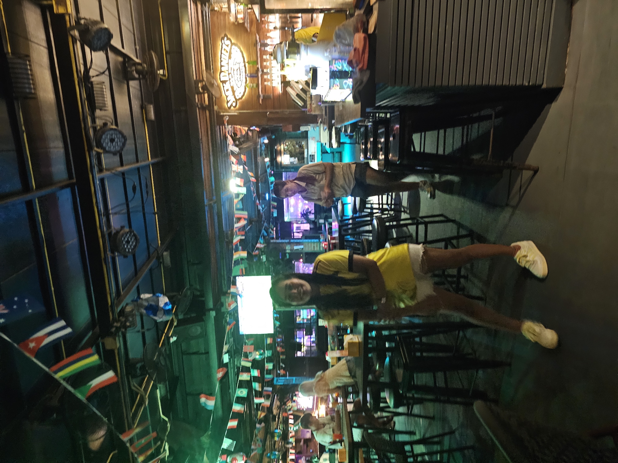 The image shows a group of people at a bar or restaurant at night in Ban Klong Haeng, Thailand. The scene is dimly lit, with neon lights and spotlights illuminating the area. In the foreground, a person wearing a yellow t-shirt and white shorts is standing, with their legs extended. In the background, other people are seated at tables, and there is a bar area with bottles and glasses. The walls are decorated with numerous small flags from different countries. A prominent sign with the text "JACK DANIEL'S" is visible behind the bar. The atmosphere appears casual and festive, possibly a gathering or a night out. The time of day is clearly nighttime, and the weather is not discernible from the image.