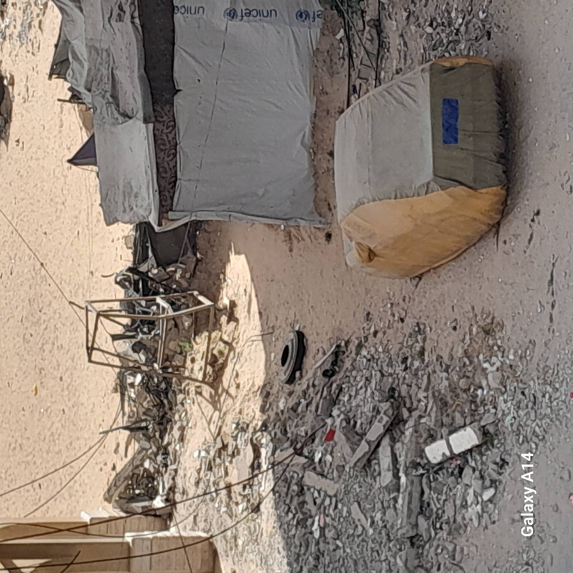 The image depicts a scene in an urban environment of Unknown, Unknown, characterized by debris and makeshift shelters. A structure covered in a white, possibly plastic material with "UNICEF" printed on it suggests a humanitarian aid setup. A metal frame and scattered rubble indicate potential damage or remnants of previous structures. A large bag lies on the ground, its contents and purpose unclear. The overall environment is dusty and appears to be in a state of disrepair or transition. There are no visible people in the image.
