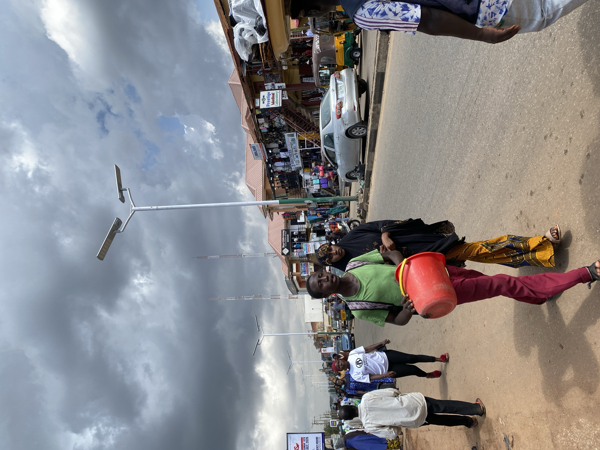 A medium shot captures a bustling street scene in Jos, Nigeria, under a heavily clouded sky. In the foreground, a young man in a green t-shirt and maroon pants carries a bright red bucket. Beside him walks a woman wearing a black hijab and a colorful patterned skirt. They are walking on a paved street lined with shops and market stalls. To the left, a group of people are walking, some on the side of the road and others on the street. Further down the street, several cars and a three-wheeled auto-rickshaw are visible. The architecture of the shops suggests a busy commercial area, with signs for various businesses, including a "Prestige Medical Laboratory." Streetlights are prominent, indicating a well-developed urban environment. The overall impression is one of everyday life unfolding in a vibrant marketplace.