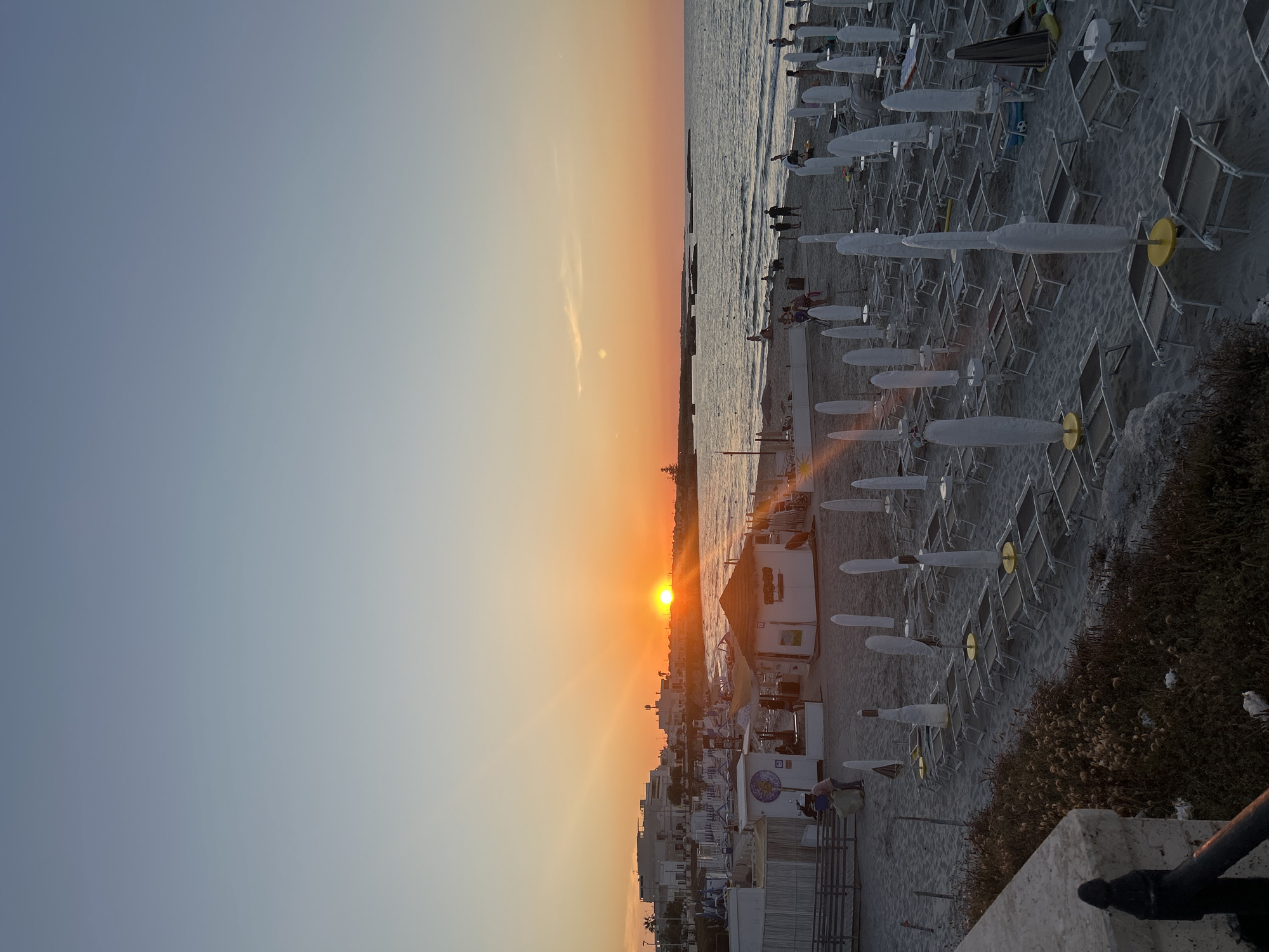 The image depicts a beach scene at sunset in San Foca, Italy. The sun is low on the horizon, casting an orange glow across the sky and sea. Numerous beach chairs and umbrellas are neatly arranged on the sand, with many of the umbrellas closed and covered. Several small structures, possibly kiosks or changing rooms, are visible on the beach. A few people are present, some walking along the shoreline and others near the structures. The water appears calm, with gentle waves lapping at the shore. Buildings line the coast in the background, suggesting a developed area. The overall impression is one of a tranquil evening on the Adriatic coast.
