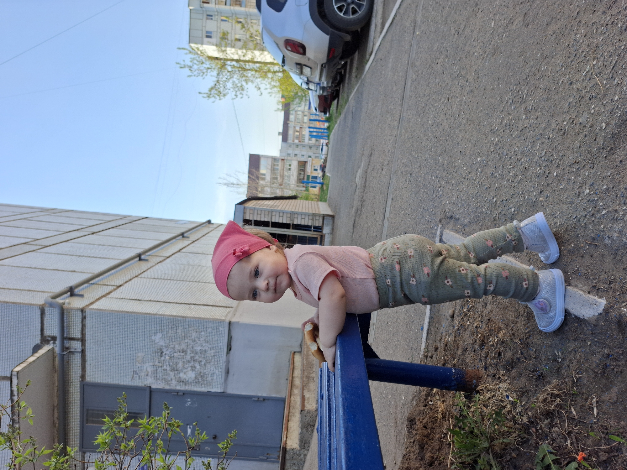 A young child stands at a blue metal fence in Chaikovsky, Russia. The child wears a pink headband, pink shirt, and patterned green pants. The child is leaning on the fence, holding a piece of bread. A gray apartment building is behind the fence to the left. A gray sidewalk is to the right. In the background are more apartment buildings, a white car, and trees. The sky is blue. The ground near the fence is dirt with some plants.