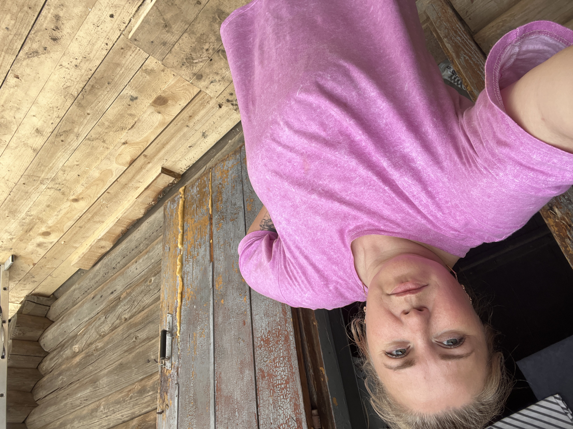 A woman is taking a selfie in front of a rustic, weathered wooden structure. She is wearing a pink t-shirt and has blonde hair. Her face is visible, and she appears to be looking directly at the camera with a neutral expression. The background is dominated by rough-hewn wooden planks, some of which are painted a peeling grey with hints of orange showing through. The wood appears old and weathered, suggesting an outdoor setting, possibly a porch or an old building. The lighting suggests it might be daytime. The setting cues, such as the aged wood and the rustic nature of the structure, point to a rural or historical location. There is no visible text in the image.