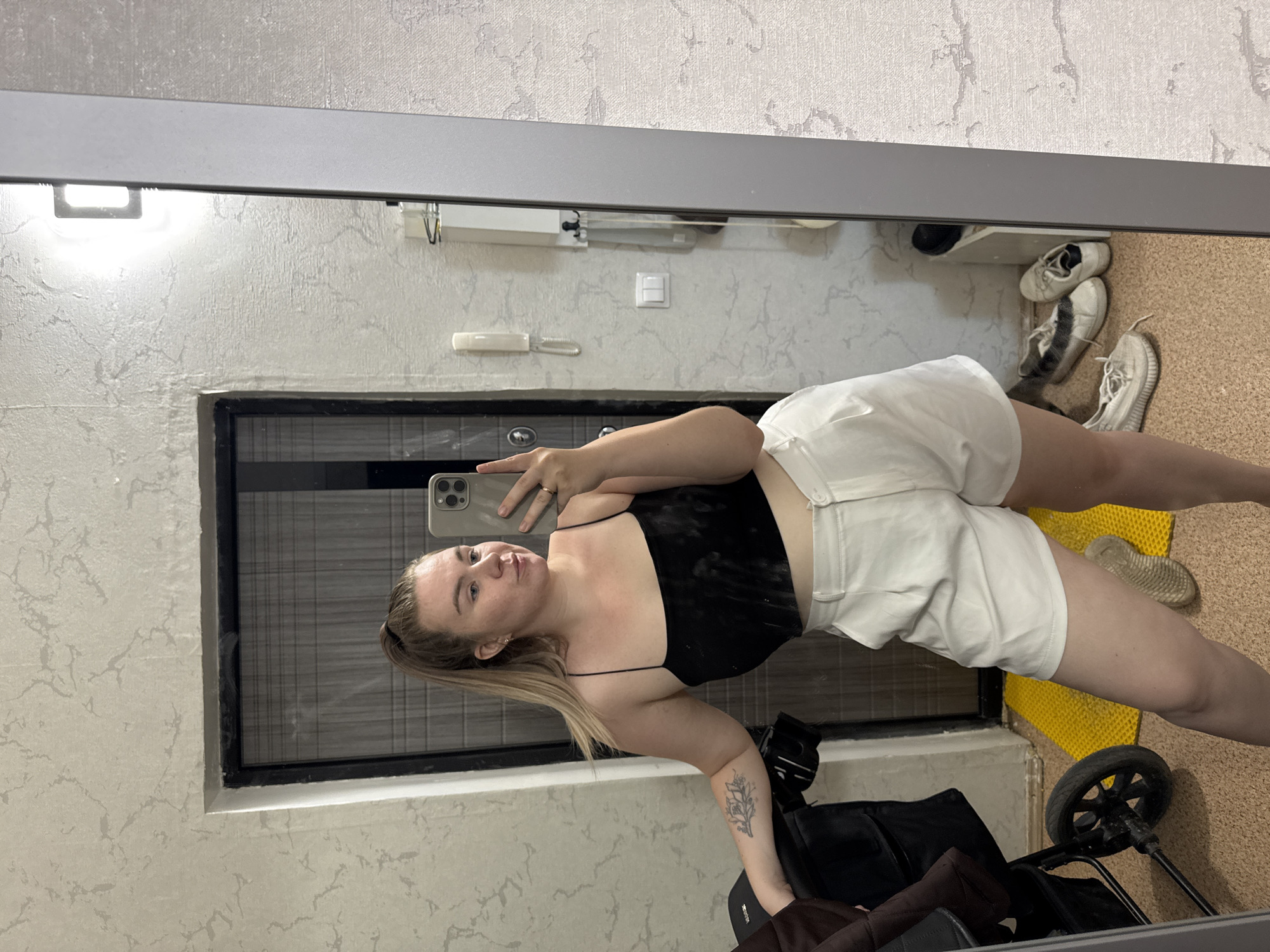 A woman is taking a selfie in a mirror inside what appears to be an apartment or home in Chaikovsky, Russia. The mirror's reflection shows a gray wall with a door and a white telephone landline receiver. She is wearing a black spaghetti-strap top and white shorts. A stroller is next to her and various shoes are organized on a rack in the background. A yellow mat can also be seen on the floor.