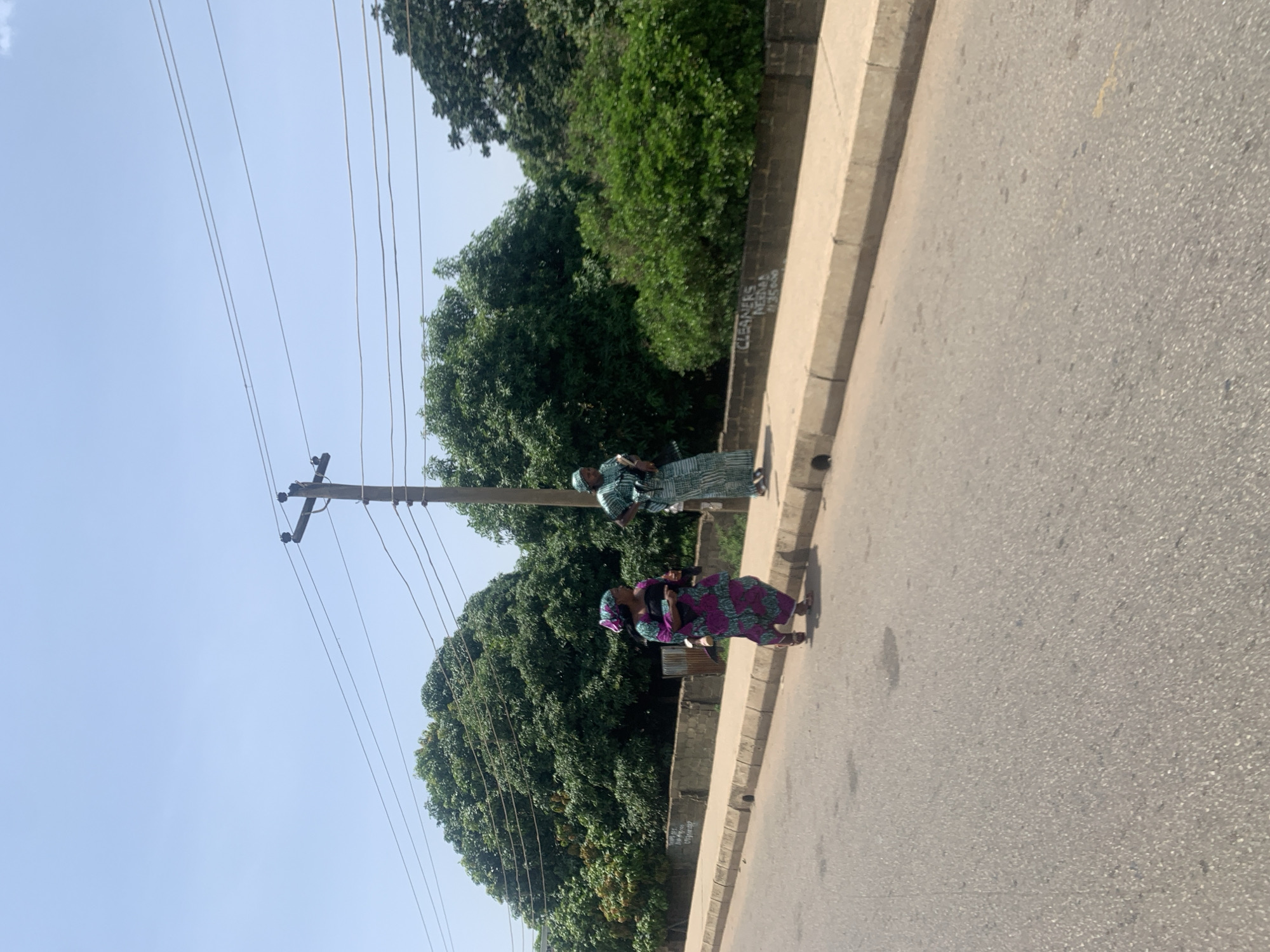 Two women are walking on the sidewalk next to a road in Jos, Nigeria. The woman on the left is wearing a pink and purple patterned dress and a matching headscarf. She is carrying a baby. The woman on the right is wearing a light blue and white striped dress and a headscarf. Both women are walking in the same direction. The road is paved and appears to be in good condition. There are trees and vegetation behind the sidewalk. A utility pole with wires is visible in the background. The sky is clear and blue. The scene appears to be in a semi-urban area.