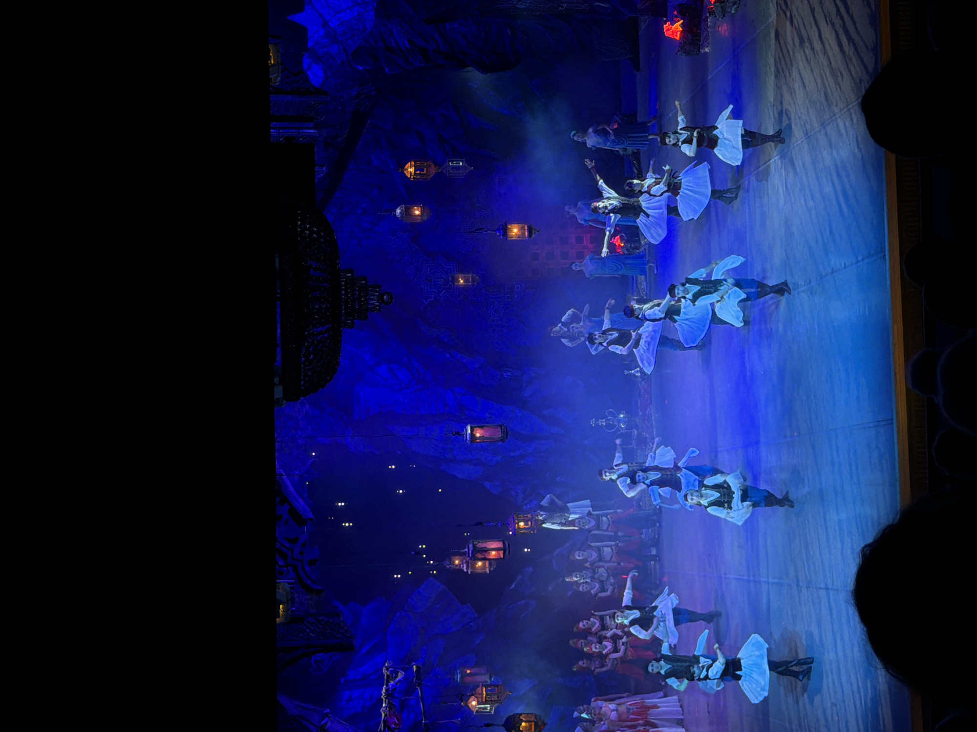 The image shows a dance performance on a stage, bathed in blue lighting. A group of dancers, wearing similar costumes of dark vests and white skirts or shirts, are performing synchronized movements. Lanterns hang above the stage, adding to the atmosphere. The backdrop is a textured wall, possibly imitating rock formations.
Although the prompt mentions the city of Izhevsk, Russia, this information is not visually discernible from the image.