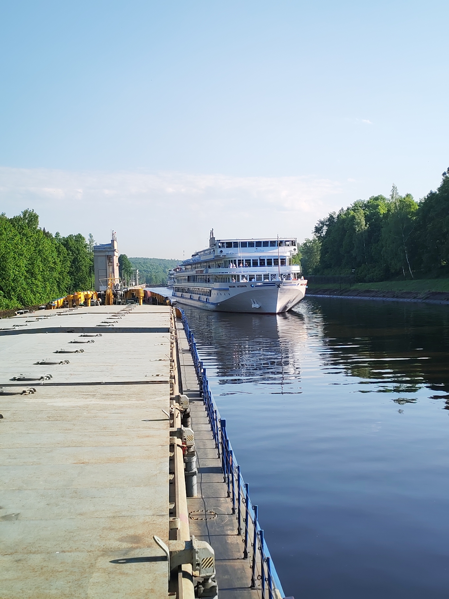 The image depicts a canal scene in Iksha, Russia, featuring a multi-deck cruise ship. The ship, white with blue trim, is moving along the canal, its reflection visible in the water. Passengers are visible on the decks.
To the left is a barge or large platform, with a concrete surface marked by rectangular panels and attachment points. Yellow mechanisms are mounted on the barge. Adjacent to the water is a blue metal safety railing.
The canal banks are lined with trees, creating a green backdrop. In the distance, a bridge-like structure with statues is visible, suggesting an elaborate system of locks and waterways. The sky is clear blue.