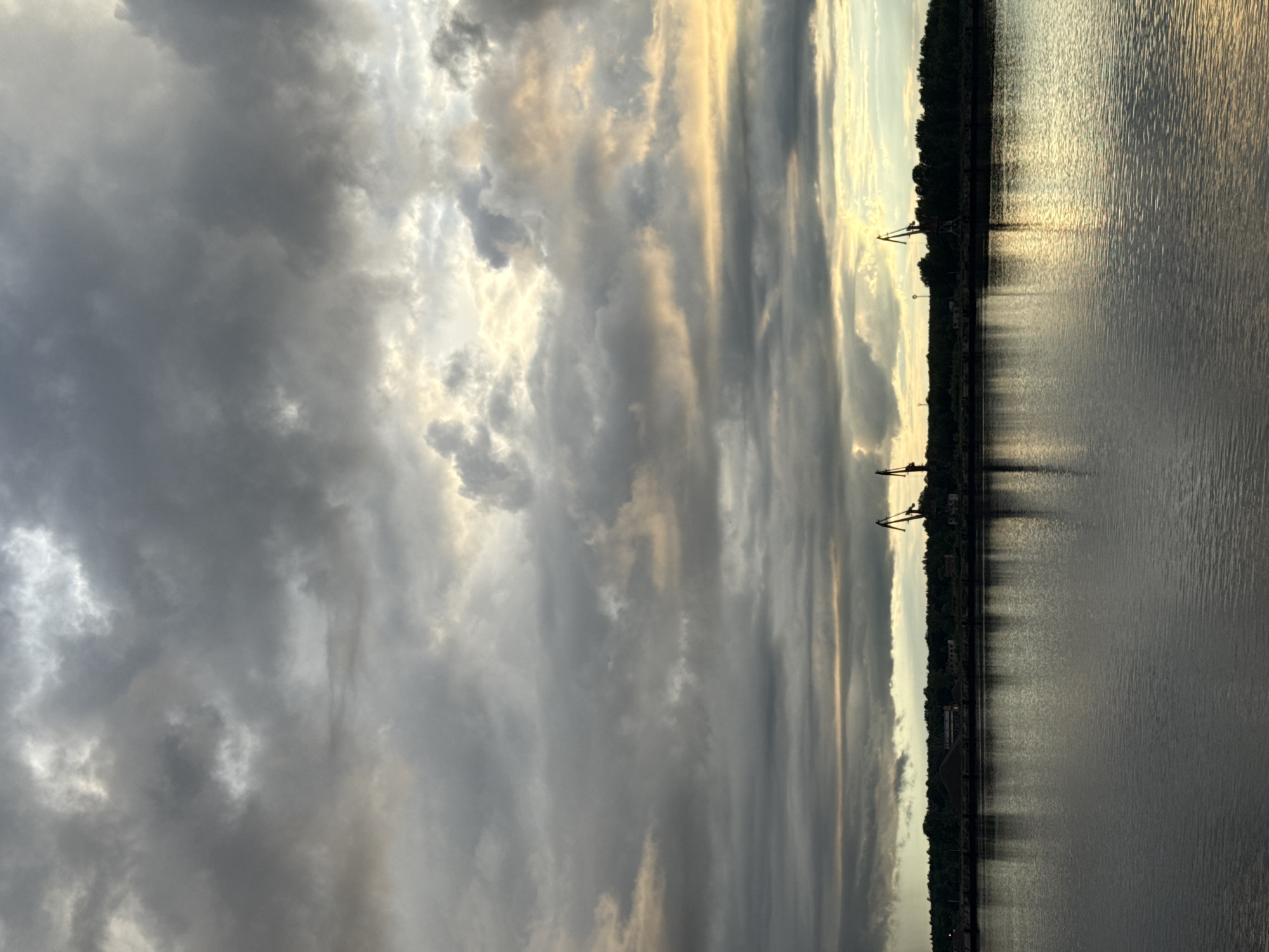 This striking image captures a waterscape under a dramatic, overcast sky, likely at either sunrise or sunset, in what appears to be an industrial river or coastal setting near Chaikovsky, Russia.
The upper portion of the frame is dominated by a vast expanse of heavy, dark grey clouds, which are thick and unevenly distributed. Glimpses of bright, golden-yellow light pierce through these clouds, illuminating their edges and creating vertical streaks of light that suggest the sun is low on the horizon or breaking through a dense cloud layer. This contrast between the dark, moody clouds and the vibrant light creates a powerful visual effect.
A dark, silhouetted shoreline stretches across the middle of the image, forming a distinct horizon line that separates the sky from the water. This landmass is largely comprised of a dense line of trees or vegetation, indicating a natural environment. Interspersed along this dark silhouette are several large, crane-like structures, typical of a port or industrial waterfront. These man-made elements stand in stark contrast to the natural beauty of the sky and water.
The lower half of the image is occupied by the water, which appears dark and reflective, mirroring the subdued tones of the cloudy sky. The golden light from above casts shimmering, vertical reflections across the water's surface, creating a textured pattern of ripples and light streaks that extend from the horizon towards the viewer. The overall mood is tranquil yet dramatic, highlighting the interplay between nature and industrial presence at a transitional time of day. No people or visible text are present in the scene.