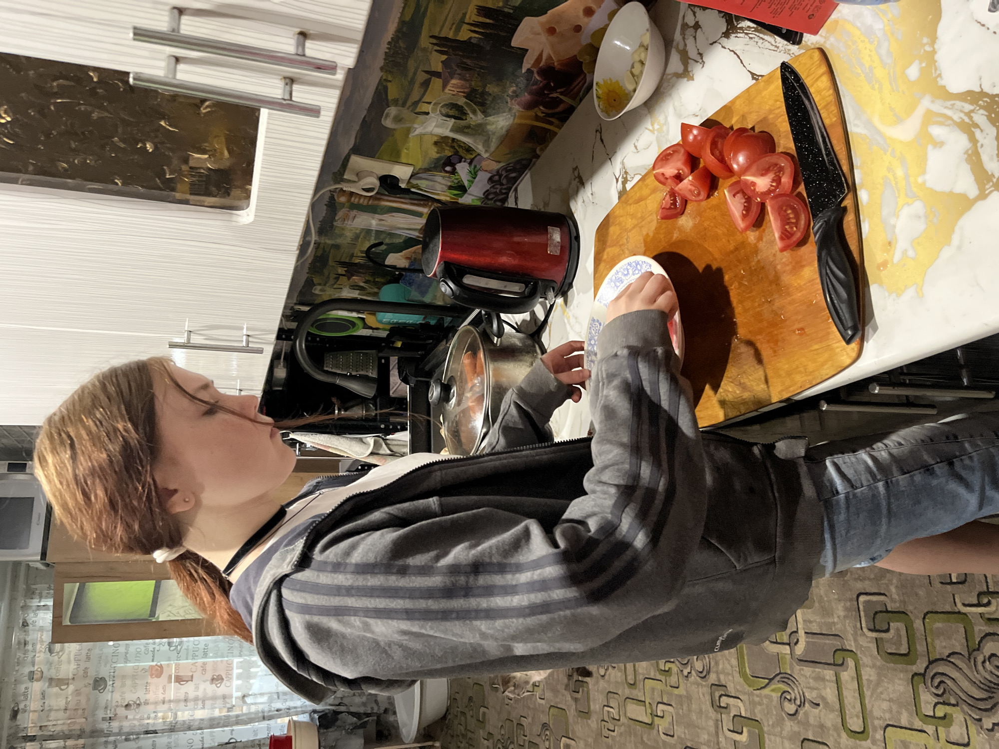 The image captures a person, likely a young woman, preparing food in a kitchen located in Vesyoloye, Russia. The photograph is oriented sideways, rotated 90 degrees counter-clockwise.
The central subject, with light brown hair tied back in a ponytail, is seen in profile. She wears a grey zip-up hoodie with three dark stripes on the sleeves and blue jeans. Her hands are holding a white plate with a blue pattern, positioned over a wooden cutting board. On the cutting board, several red tomatoes are neatly sliced. A black-handled kitchen knife with a speckled black blade rests beside them.
The kitchen counter features a white surface with distinctive golden, marble-like patterns. To the left of the cutting board, a red electric kettle is visible, and further back, a cooking pot with a glass lid is on a stovetop. A white bowl containing what appears to be garlic cloves and a yellow substance is also on the counter. The backsplash behind the counter displays a scenic painting of a countryside. White kitchen cabinets with silver handles are mounted above.
The floor has a textured pattern of interconnected green and brown geometric shapes. In the background, on a glass surface, various coffee-related terms are repeatedly printed, including "cafe latte," "CAPPUCCINO," "coffee," and "MOCHA LATTE." The lighting suggests an indoor setting, possibly in the evening. The scene depicts a routine domestic activity of meal preparation.