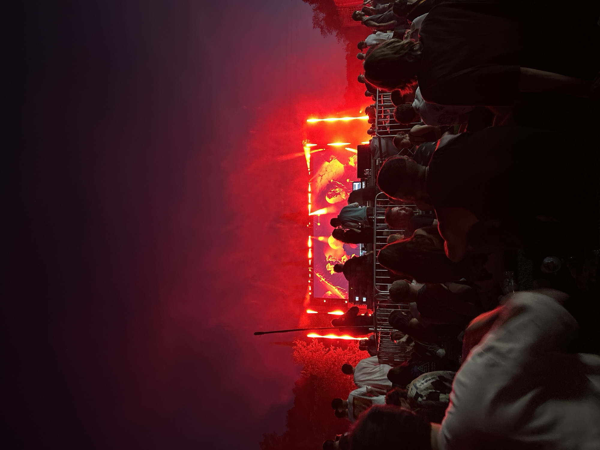The image depicts an outdoor music festival at night in Castellbisbal, Spain. A crowd of people is gathered, facing a brightly lit stage. The stage is adorned with vibrant red and orange lights, creating a dramatic atmosphere. Several individuals are visible on the stage, appearing to be performers or DJs, interacting with equipment. The audience is densely packed, with many silhouetted figures facing the stage. Some people are seated on the ground, while others are standing. The sky above is dark, with the bright stage lights illuminating the surrounding area and casting a warm glow on the crowd and trees. The overall scene suggests an energetic and engaging concert experience.